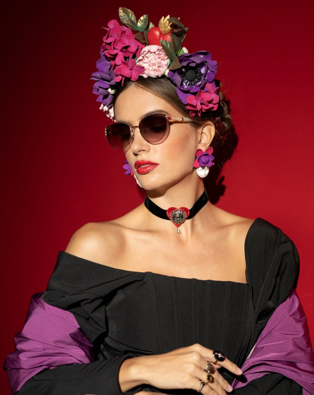 A woman with floral headwear and ultra-lightweight Frida Brush sunglasses by For Art&#39;s Sake® poses against a red background. She wears a black off-the-shoulder dress with purple sleeves, a black choker with a red heart pendant, and bold red lipstick. Her gold-plated earrings and hairstyle complement the vibrant flowers.