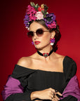 A woman with floral headwear and ultra-lightweight Frida Brush sunglasses by For Art's Sake® poses against a red background. She wears a black off-the-shoulder dress with purple sleeves, a black choker with a red heart pendant, and bold red lipstick. Her gold-plated earrings and hairstyle complement the vibrant flowers.