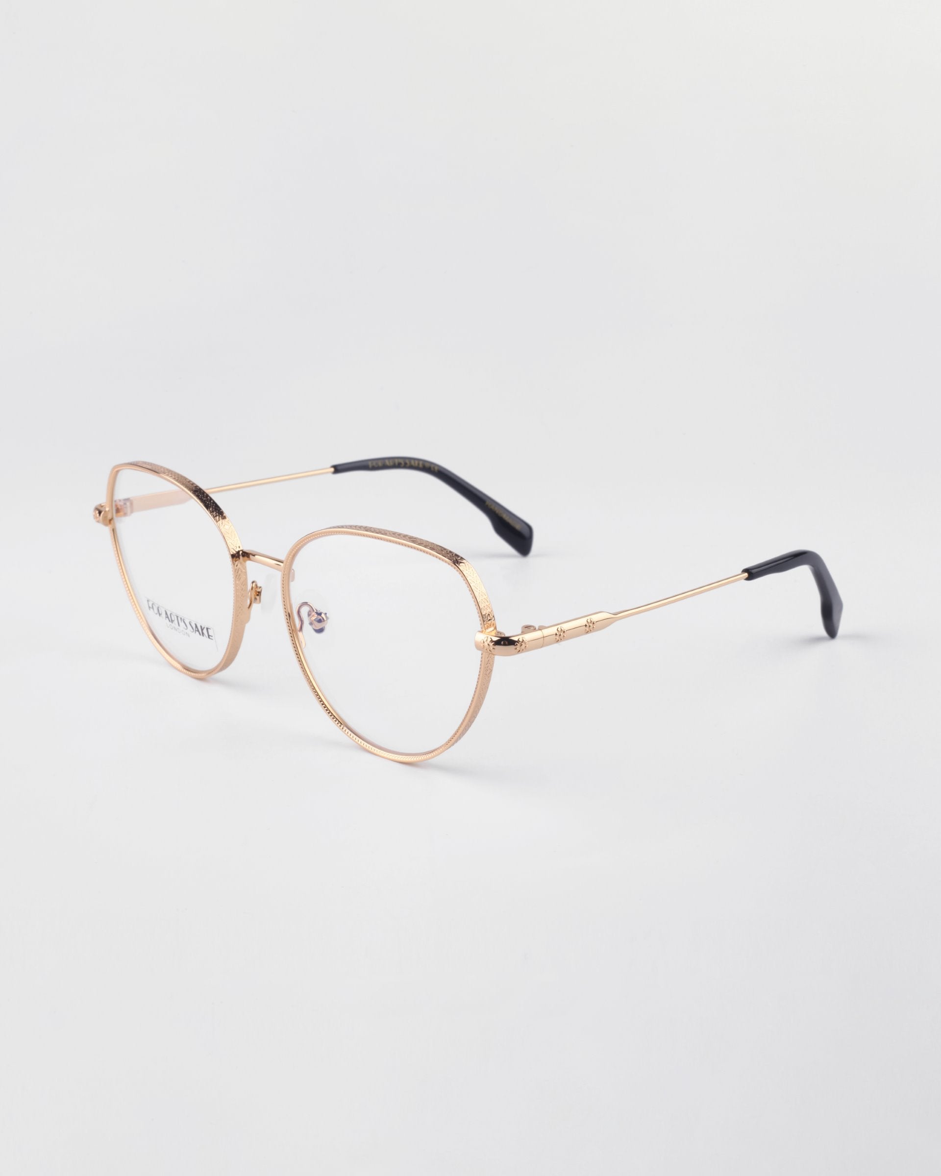A pair of Frida Floral eyeglasses by For Art&#39;s Sake® with round lenses and black temple tips rests on a white surface. The glasses have a sleek, minimalist design with thin metal frames, a subtle bridge, and prescription lenses.