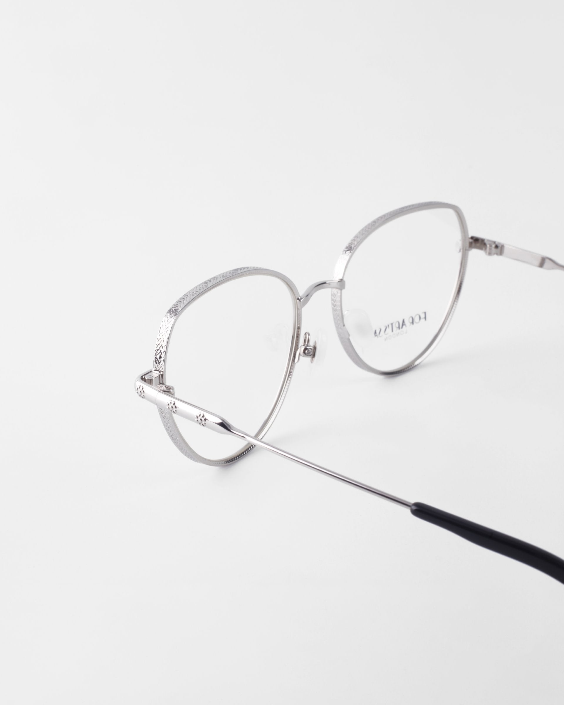 A pair of Frida Floral by For Art&#39;s Sake® rests on a white surface. The straight arm of the glasses extends towards the camera, showcasing a sleek design with a black tip. The brand name is faintly visible on the lens, complemented by an 18-karat gold-plated frame.