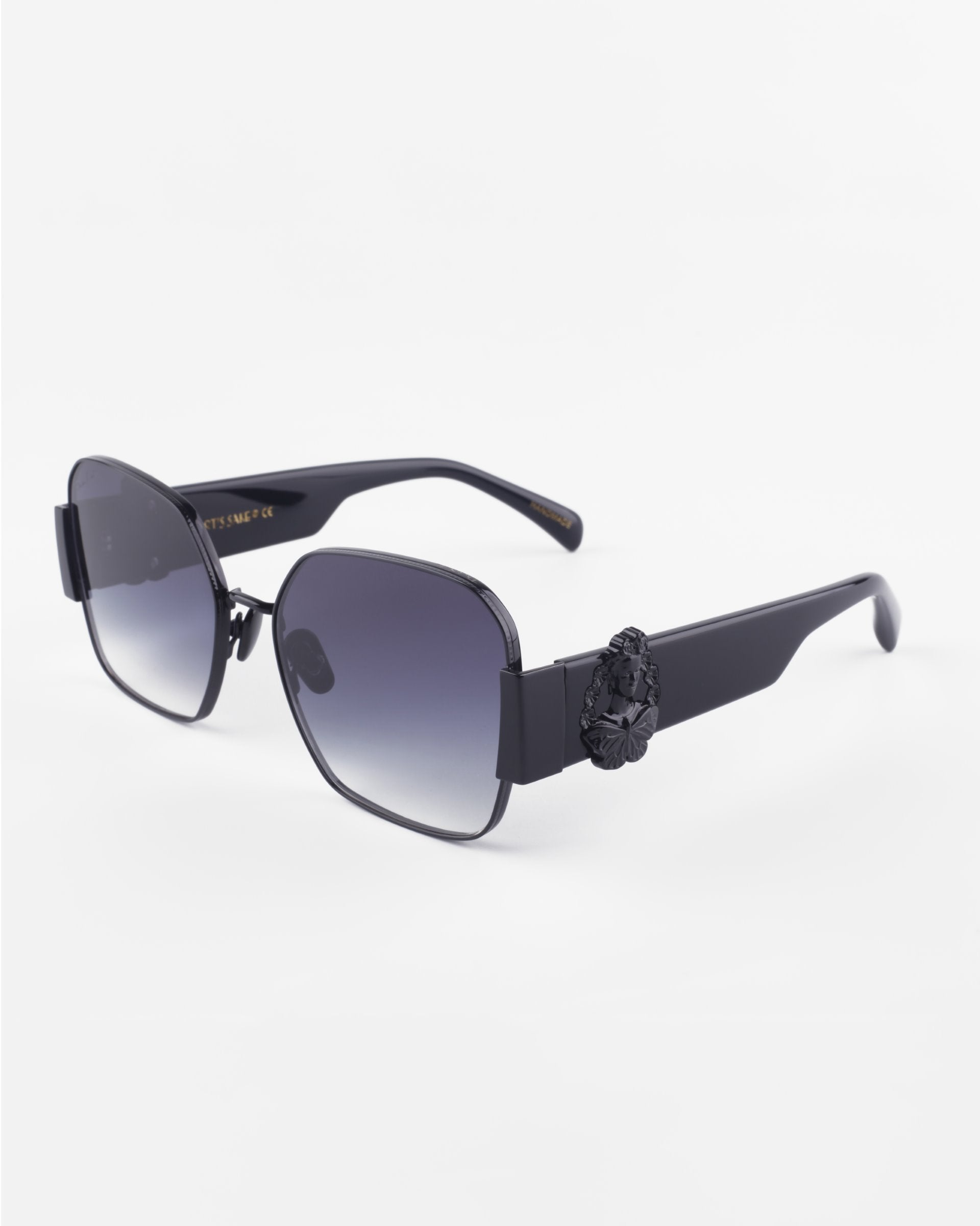 A pair of stylish, oversized black sunglasses with square lenses that have a gradient tint from dark to light. Featuring ultra-lightweight nylon lenses and 100% UVA &amp; UVB protection, the Frida Mask by For Art&#39;s Sake® also boasts a decorative black piece on the gold-plated stainless steel hinges against a white background.
