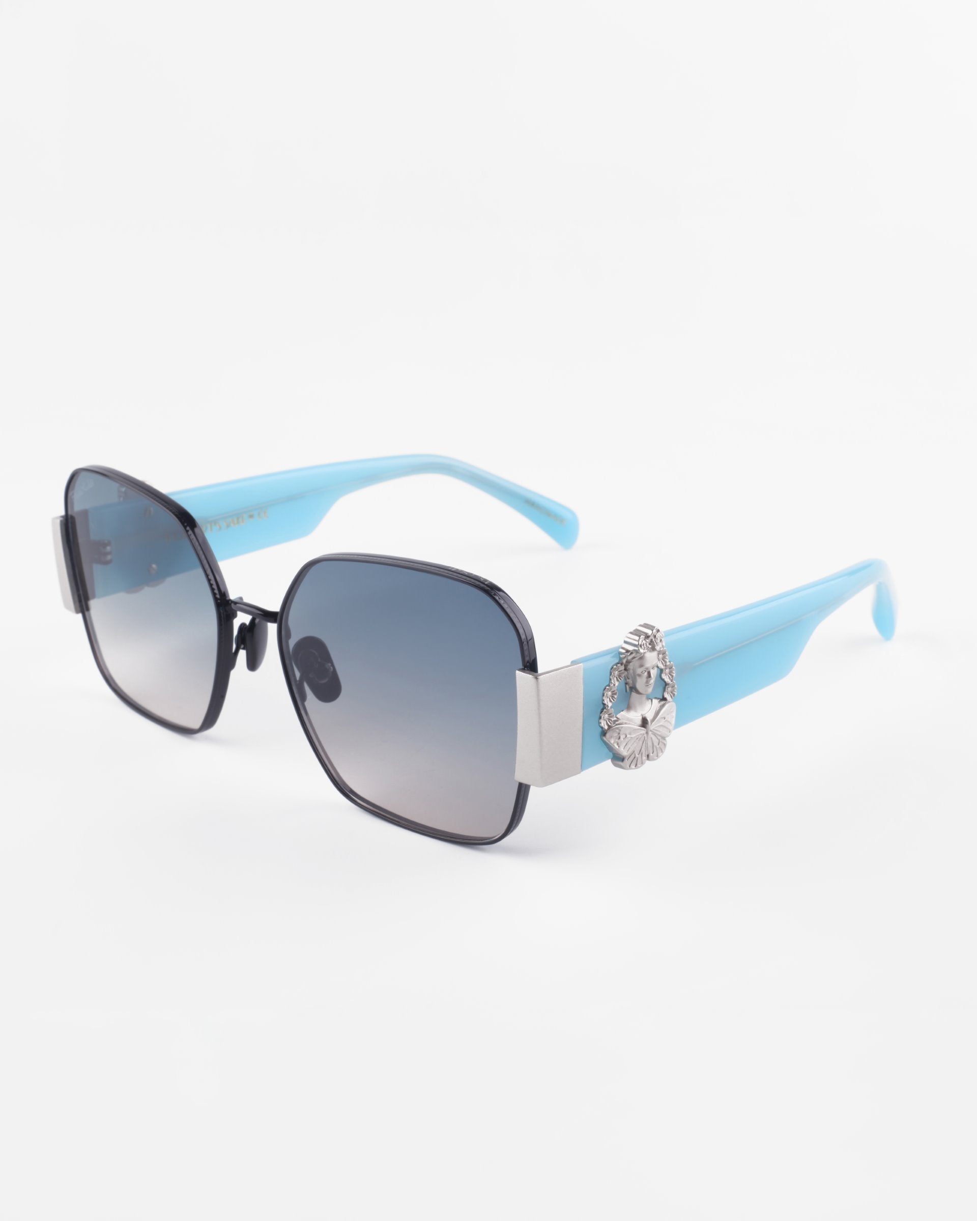 Introducing the Frida Mask by For Art&#39;s Sake®: A pair of rectangular sunglasses featuring black frames and light blue temples. The temples have a silver decorative element with a circular emblem near the hinge. The ultra-lightweight Nylon lenses offer gradient shading, transitioning from dark at the top to lighter at the bottom, with 100% UVA &amp; UVB protection.