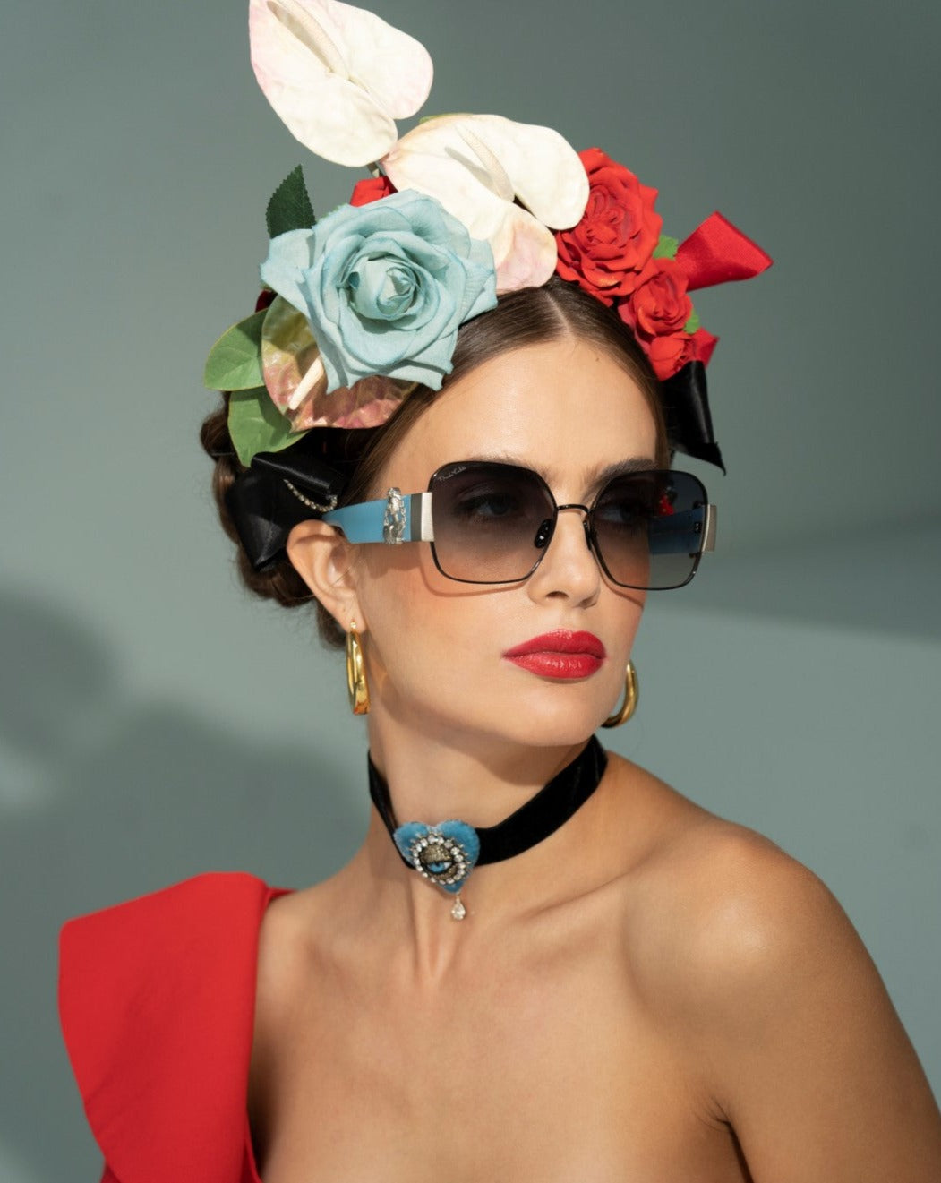 A woman wearing oversized For Art&#39;s Sake® Frida Mask with ultra-lightweight Nylon lenses, a black choker with a blue brooch, and a red off-the-shoulder outfit. She has red lipstick and a floral headpiece adorned with blue, white, and red flowers. The background is plain and grey.