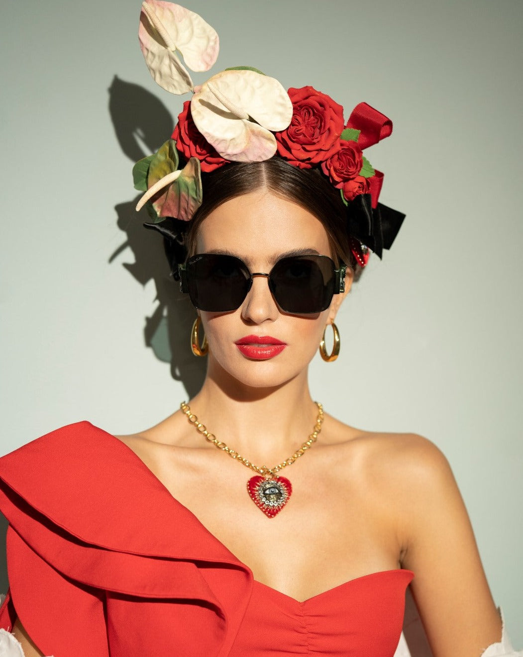 A woman wearing oversized black sunglasses with 100% UVA &amp; UVB protection, a red off-the-shoulder dress with a large ruffle, and large gold hoop earrings. Her hair is adorned with red roses and white flowers, and she sports red lipstick and a gold-plated stainless steel necklace with a heart-shaped pendant (**Frida Mask by For Art&#39;s Sake®**).