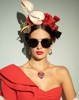 A woman wearing oversized black sunglasses with 100% UVA & UVB protection, a red off-the-shoulder dress with a large ruffle, and large gold hoop earrings. Her hair is adorned with red roses and white flowers, and she sports red lipstick and a gold-plated stainless steel necklace with a heart-shaped pendant (**Frida Mask by For Art's Sake®**).