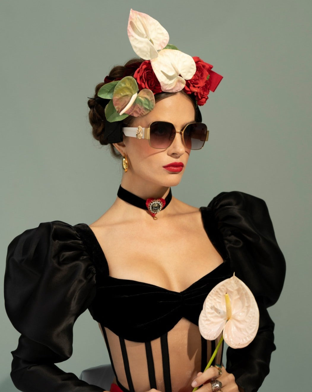 A woman in stylish, modern attire poses against a plain background. She wears oversized, ultra-lightweight For Art&#39;s Sake® Frida Mask with 100% UVA &amp; UVB protection, a black dress with puffed sleeves, a floral headpiece, and a choker necklace. Holding light pink flowers, her look is sophisticated and fashion-forward.