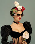 A woman in stylish, modern attire poses against a plain background. She wears oversized, ultra-lightweight For Art's Sake® Frida Mask with 100% UVA & UVB protection, a black dress with puffed sleeves, a floral headpiece, and a choker necklace. Holding light pink flowers, her look is sophisticated and fashion-forward.