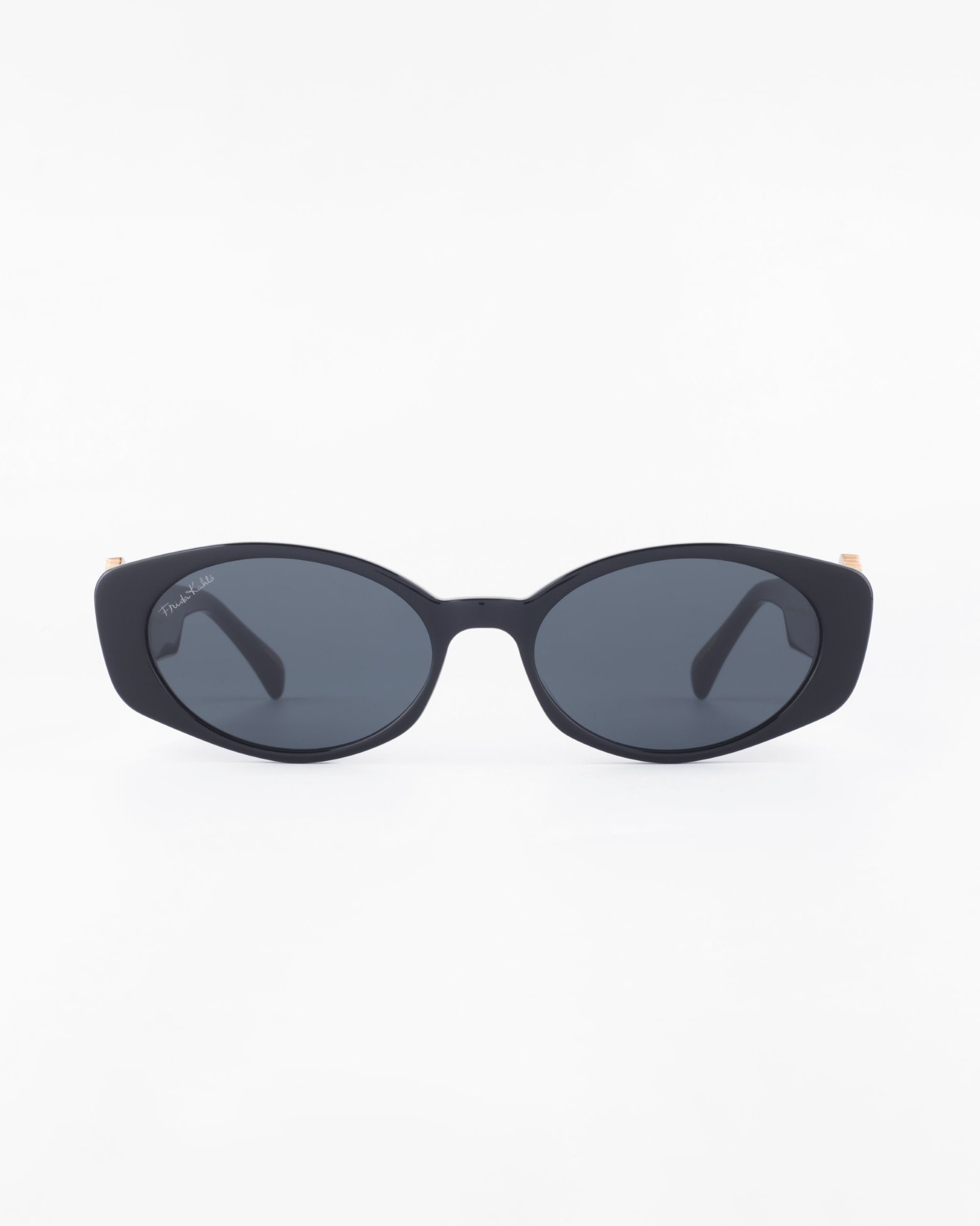A pair of black oval-shaped Frida Portrait sunglasses by For Art's Sake® with dark tinted, ultra-lightweight nylon lenses is centered on a white background. Both arms of the sunglasses are folded, and they offer 100% UVA & UVB protection.