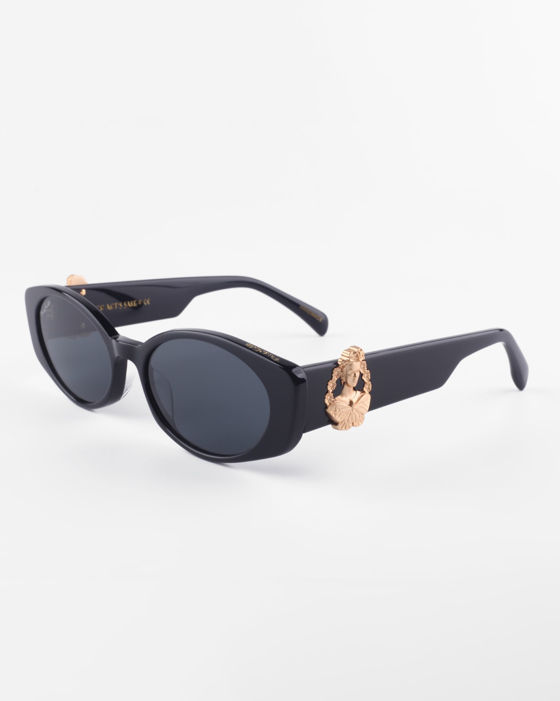 A pair of black, oval-shaped Frida Portrait sunglasses with dark lenses by For Art&#39;s Sake®. The sunglasses feature an 18-karat gold-plated decorative element on the temples, resembling a stylized, ornate design. The ultra-lightweight Nylon lenses offer 100% UVA &amp; UVB protection. The arms are wide and the frame is glossy. The background is plain white.