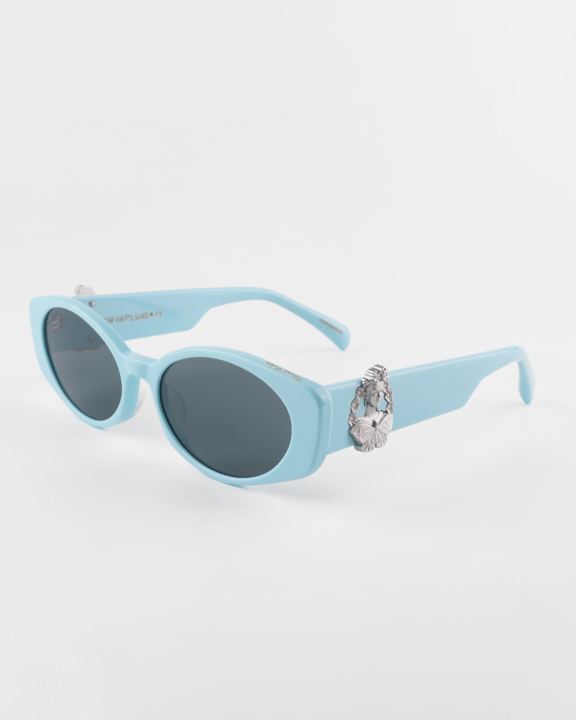 A pair of light blue, oval-shaped Frida Portrait sunglasses by For Art&#39;s Sake® with ultra-lightweight Nylon lenses that provide 100% UVA &amp; UVB protection. The frame features decorative silver embellishments on the sides, adding a touch of uniqueness to the design. The background is plain white, highlighting the stylish eyewear.