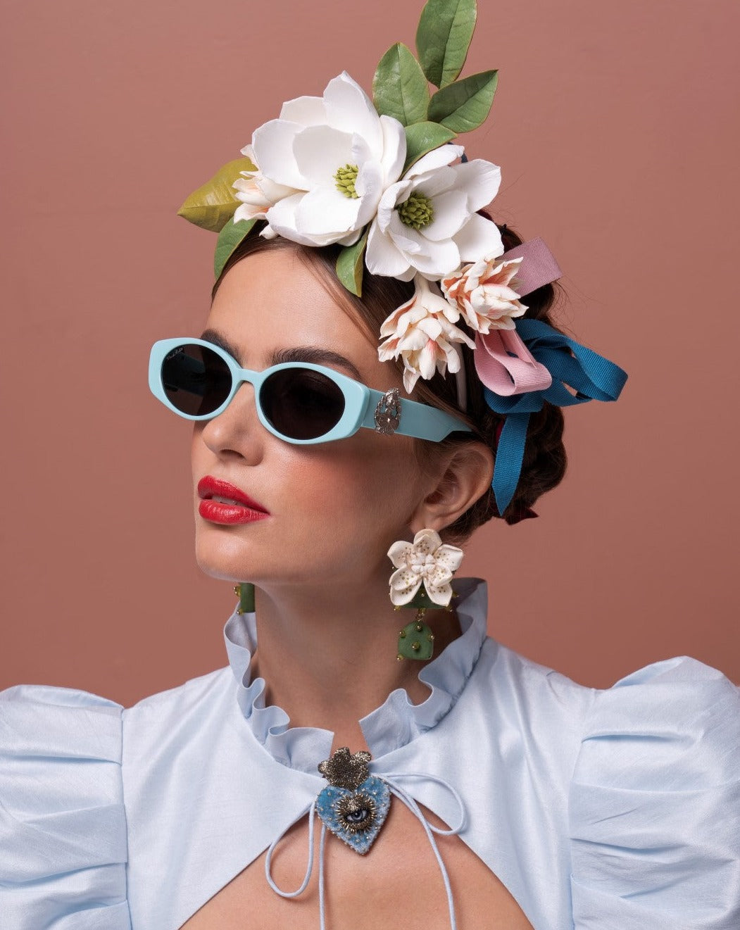 A woman wearing Frida Portrait sunglasses by For Art&#39;s Sake® with ultra-lightweight nylon lenses and a light blue dress with a ruffled neckline. She has red lipstick and is adorned with a large floral headpiece and dangling earrings. The backdrop is a warm, muted shade of pink.