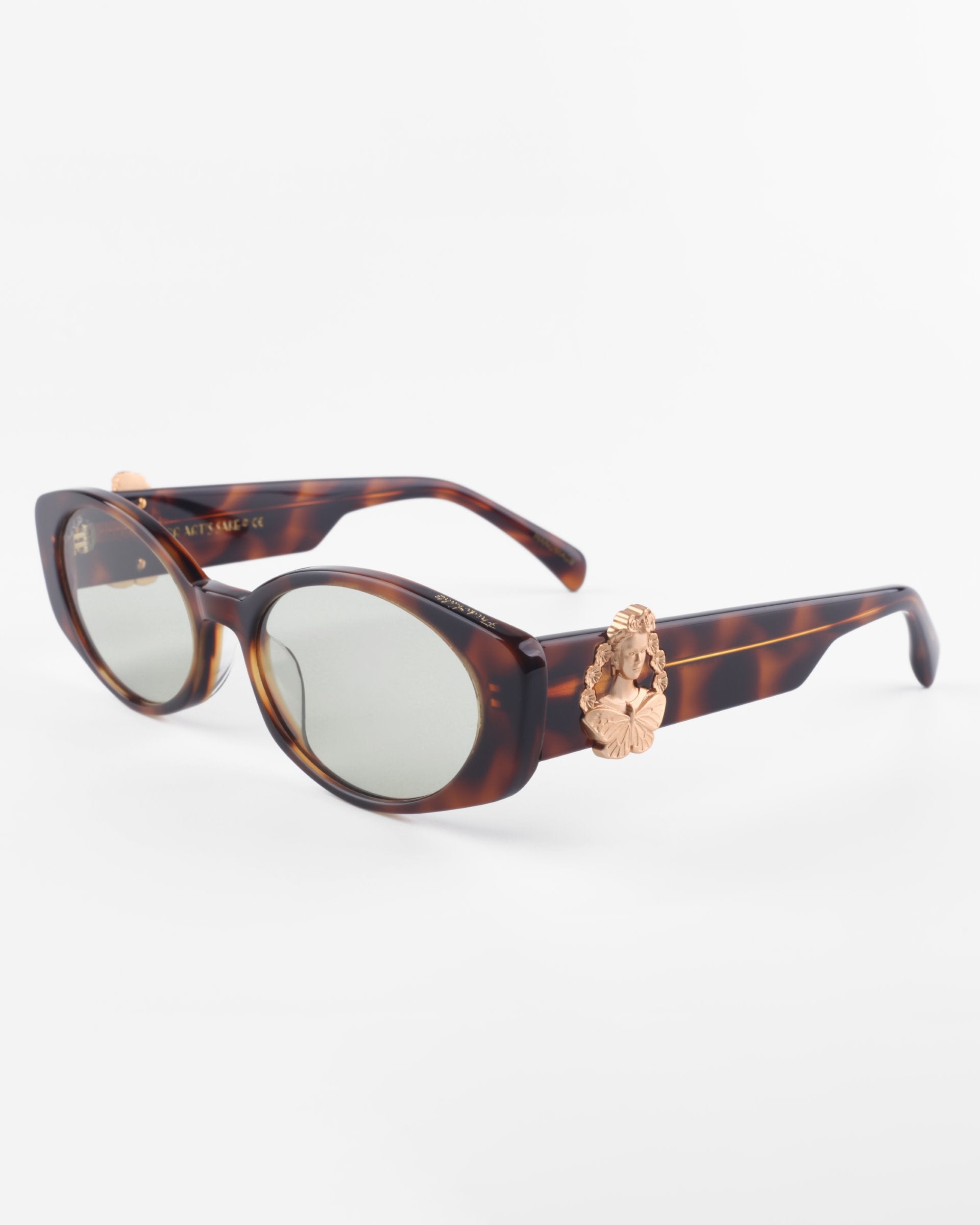 A pair of stylish Frida Portrait sunglasses by For Art&#39;s Sake® with oval lenses and tortoiseshell frames. The sunglasses feature decorative 18-karat gold-plated lion head embellishments on the temples near the hinges. The ultra-lightweight nylon lenses offer 100% UVA &amp; UVB protection and are tinted in a light gray shade. The background is plain white.