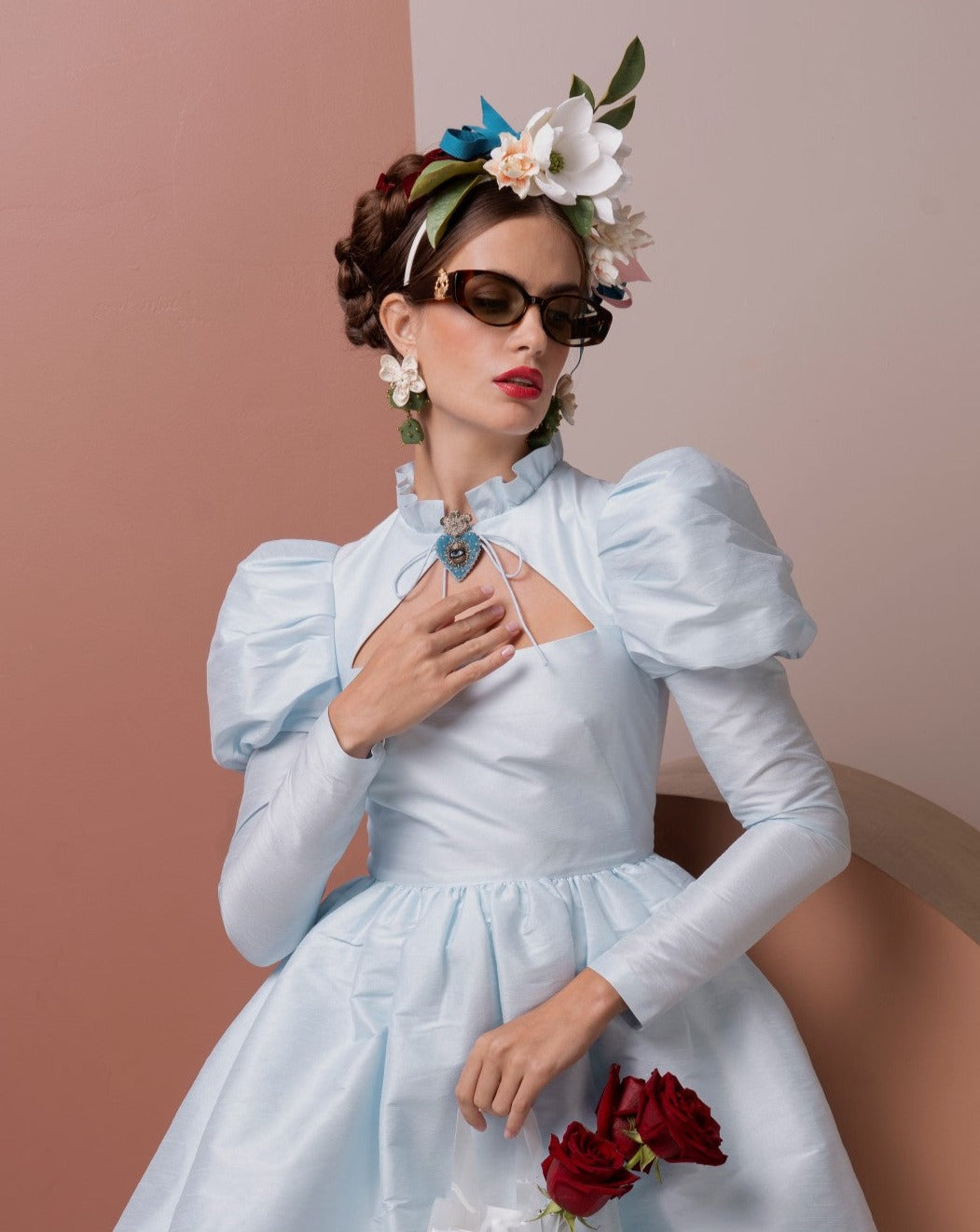 A woman dressed in a light blue vintage-style dress with puffed sleeves poses against a color-blocked background. She wears floral headgear, ultra-lightweight For Art&#39;s Sake® Frida Portrait sunglasses with 100% UVA &amp; UVB protection, drop earrings, and holds red roses in one hand while touching her chest with the other.