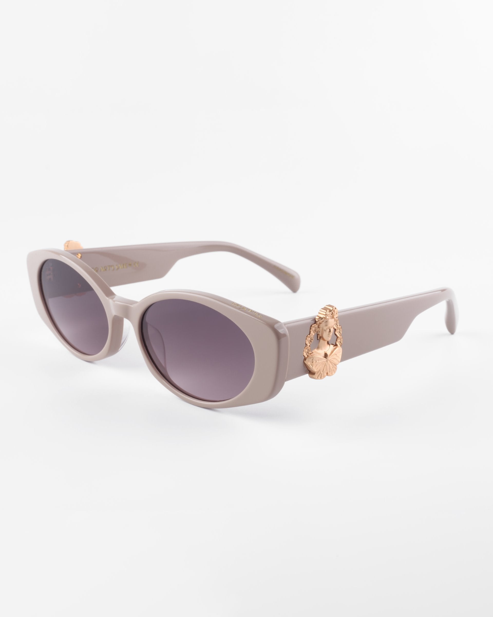 Oval-shaped, muted lavender Frida Portrait sunglasses by For Art&#39;s Sake® with ultra-lightweight nylon lenses offering 100% UVA &amp; UVB protection. The frame features an ornate, 18-karat gold-plated decorative element on the temples near the hinges. The background is a plain white surface.