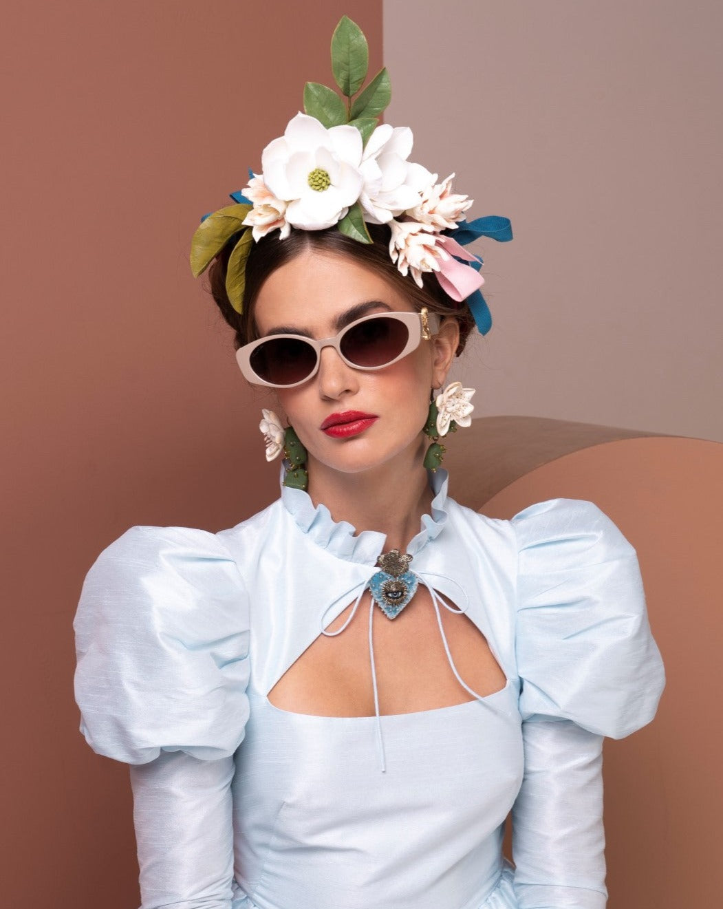 A woman wearing a light blue dress with puffed sleeves, white Frida Portrait by For Art&#39;s Sake® sunglasses with ultra-lightweight nylon lenses, and flower earrings poses confidently. She has a floral headpiece adorned with various flowers and leaves atop her dark hair. The background features earthy tones, enhancing her radiant look.