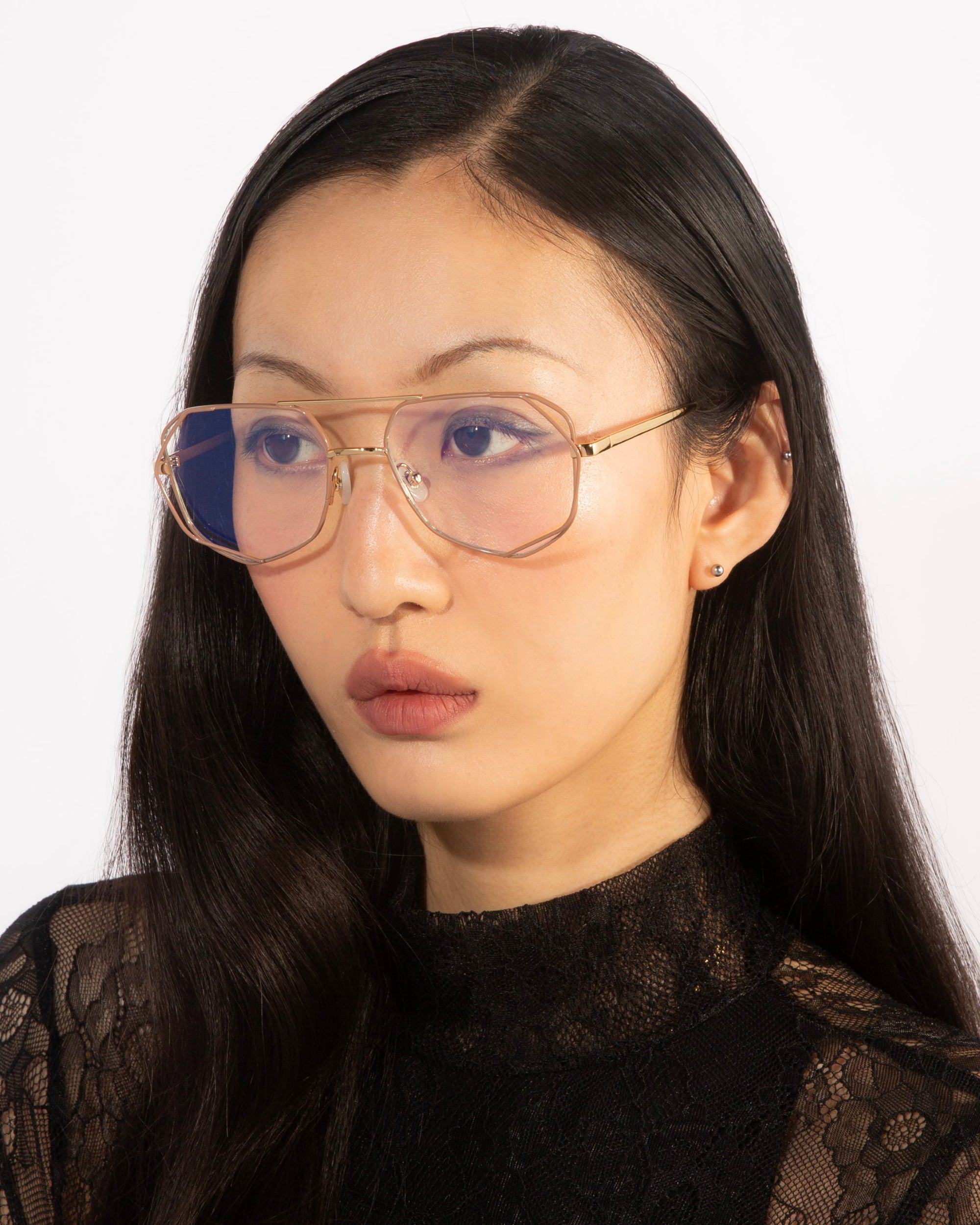 A woman with long, dark hair wearing the Genius Two glasses by For Art's Sake®, which are large and 18-karat gold-plated with a blue light filter, and a black lace top is looking slightly away from the camera against a plain white background.