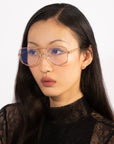 A woman with long, dark hair wearing the Genius Two glasses by For Art's Sake®, which are large and 18-karat gold-plated with a blue light filter, and a black lace top is looking slightly away from the camera against a plain white background.