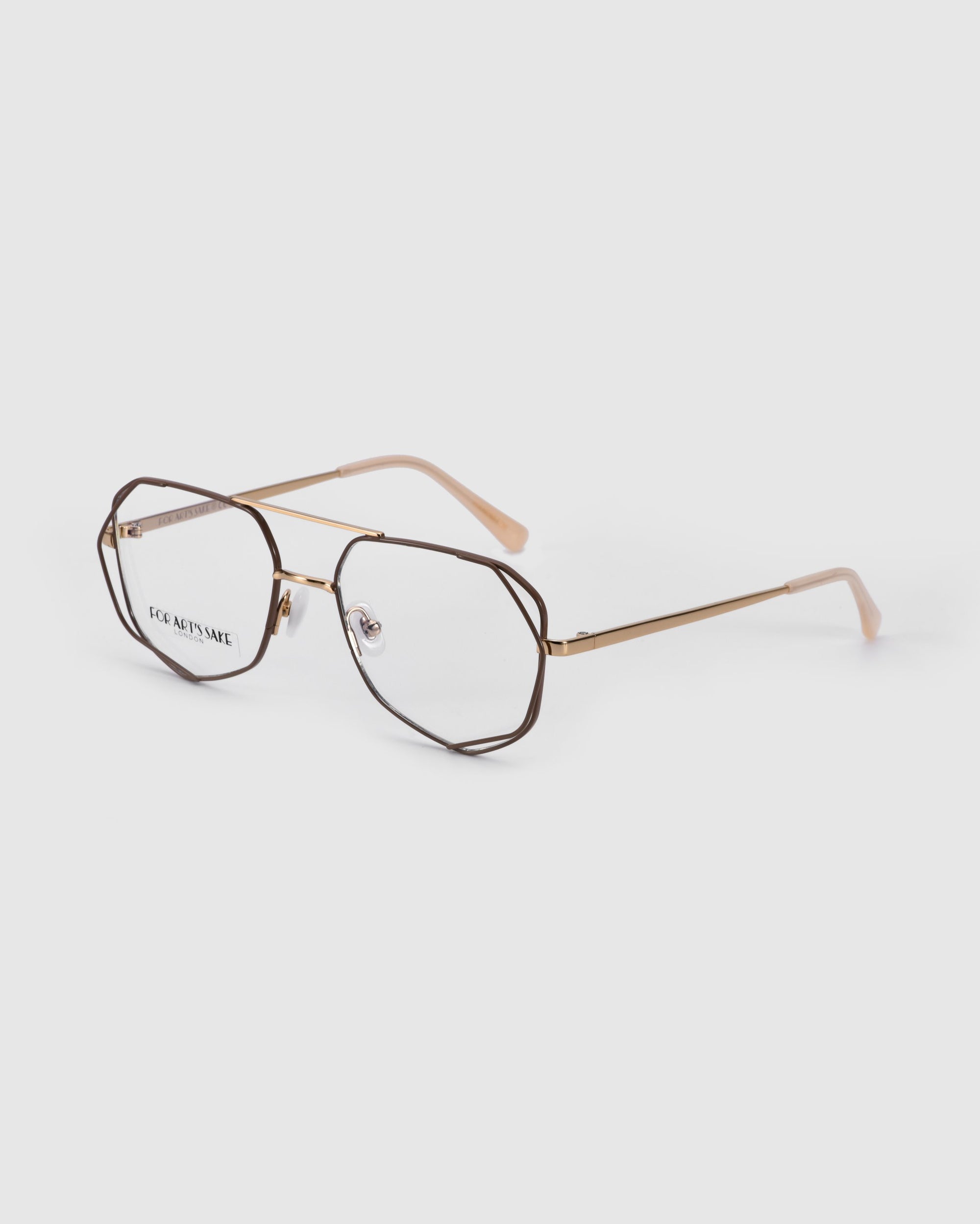 Introducing the Genius Two by For Art&#39;s Sake®: a pair of geometric eyeglasses with thin, metallic gold frames and clear lenses featuring a blue light filter. The temple tips are coated in a nude or light peach-colored material. The glasses rest angled on a white surface.