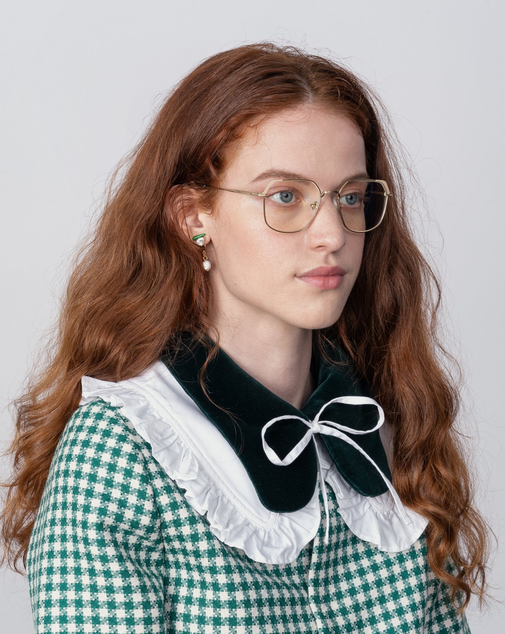 A young woman with long, wavy red hair is wearing Godiva gold-framed glasses with a blue light filter from For Art's Sake® and a green and white houndstooth dress with a large black and white ruffled collar. She is looking slightly to the side against a plain background.