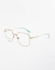 A pair of eyeglasses with gold wire frames and rectangular lenses, featuring mint green accents on the temples and end tips of the arms. These stylish glasses, named Godiva by For Art's Sake®, also come with optional prescription lenses. Displayed against a white background, they elegantly blend fashion with functionality.