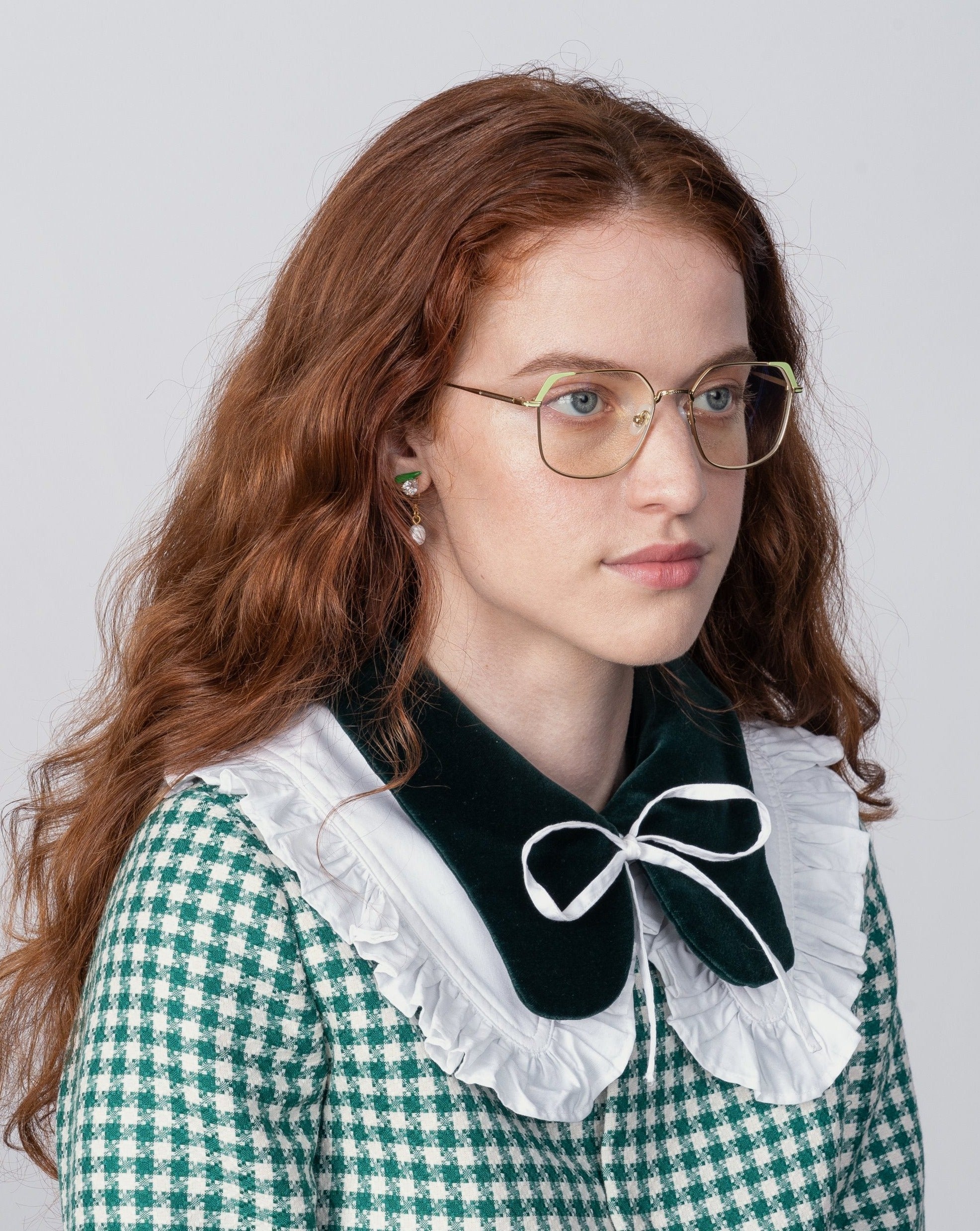 A person with long, wavy, red hair and wearing glasses with prescription lenses from For Art's Sake® looks to the side. They are dressed in a green and white checkered outfit with a large black collar adorned with a white ruffle and bow. The background is plain and light-colored.
