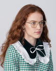 A person with long, wavy, red hair and wearing glasses with prescription lenses from For Art's Sake® looks to the side. They are dressed in a green and white checkered outfit with a large black collar adorned with a white ruffle and bow. The background is plain and light-colored.