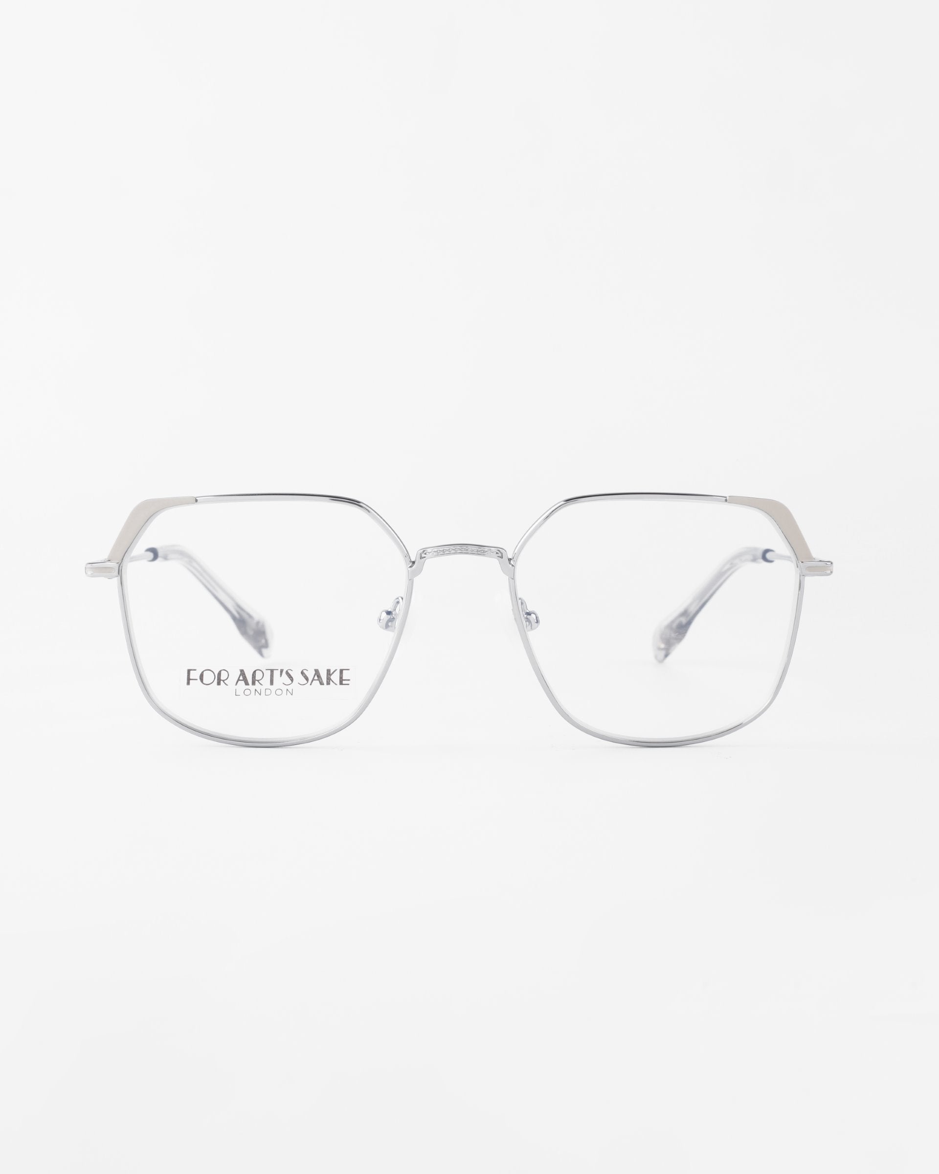 A pair of Godiva eyeglasses from For Art&#39;s Sake®, with a geometric design, featuring thin, angular metal temples and clear lenses with blue light filter. The phrase &quot;FOR ART&#39;S SAKE LONDON&quot; is printed on the left lens. The glasses are displayed against a white background.