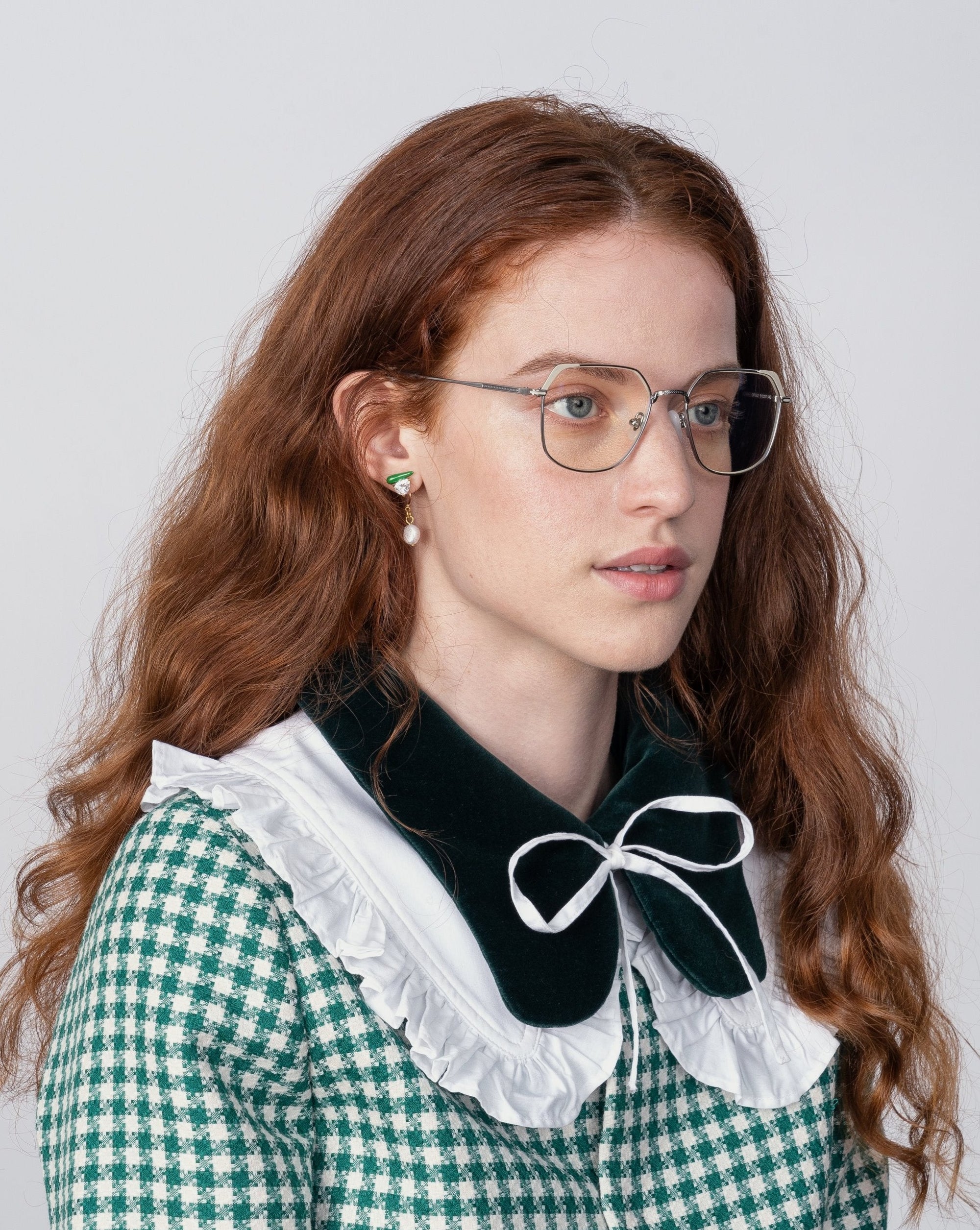 A young woman with long, wavy red hair, wearing glasses with prescription lenses and a green and white checkered dress with a large, white, ruffled collar adorned with a black bow. She has green gemstone earrings and is looking slightly off to the side against a plain, light background while wearing For Art&#39;s Sake® Godiva eyeglasses.