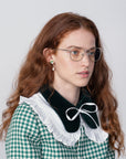 A young woman with long, wavy red hair, wearing glasses with prescription lenses and a green and white checkered dress with a large, white, ruffled collar adorned with a black bow. She has green gemstone earrings and is looking slightly off to the side against a plain, light background while wearing For Art's Sake® Godiva eyeglasses.