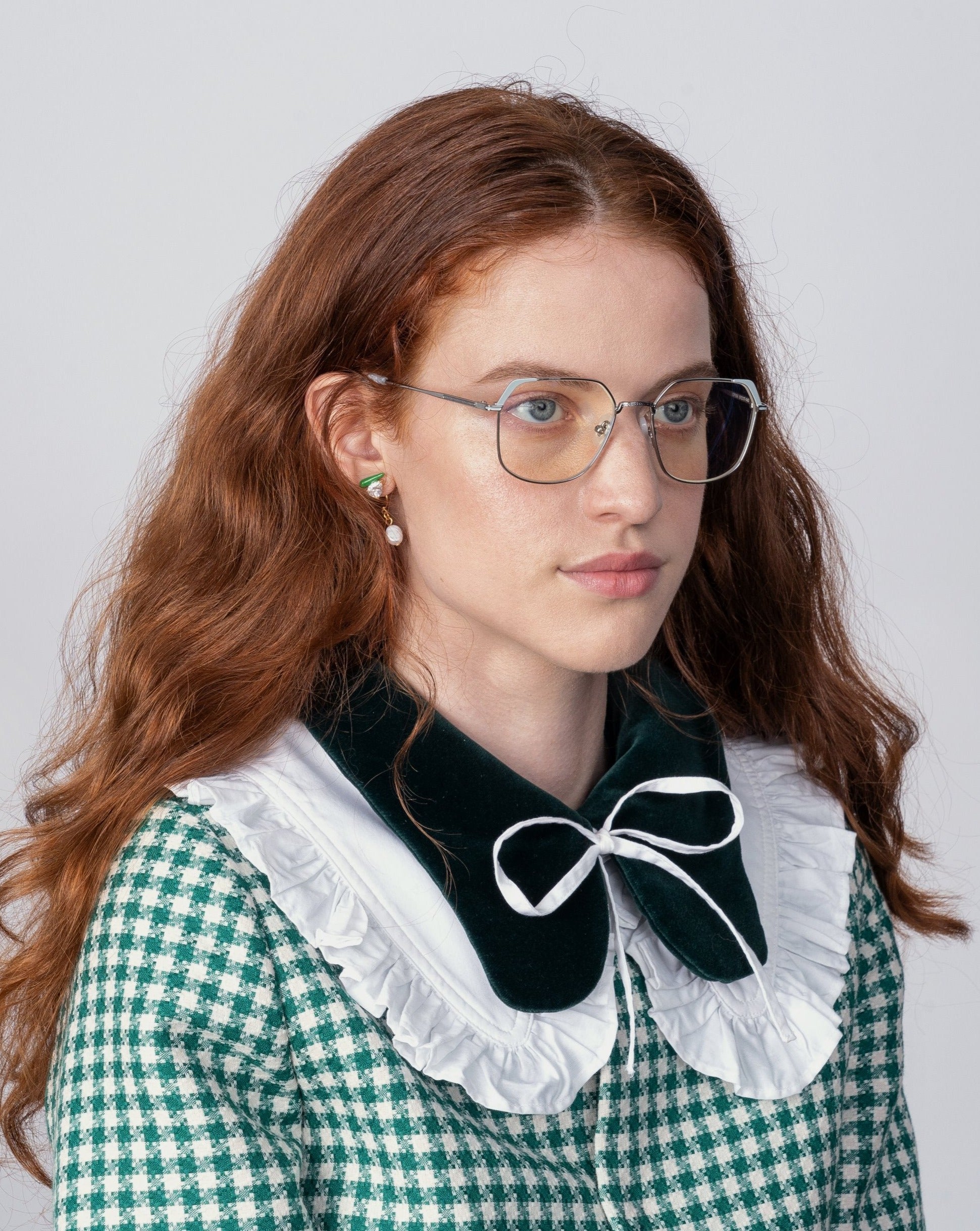 A woman with long, wavy red hair wearing Godiva by For Art&#39;s Sake® eyeglasses with prescription lenses and a blue light filter is seen from the shoulders up. She is dressed in a green and white checkered outfit featuring a dark velvet collar with a white ruffled edge and a bow tied at her neck. The background is plain and light-colored.