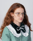 A woman with long, wavy red hair wearing Godiva by For Art's Sake® eyeglasses with prescription lenses and a blue light filter is seen from the shoulders up. She is dressed in a green and white checkered outfit featuring a dark velvet collar with a white ruffled edge and a bow tied at her neck. The background is plain and light-colored.