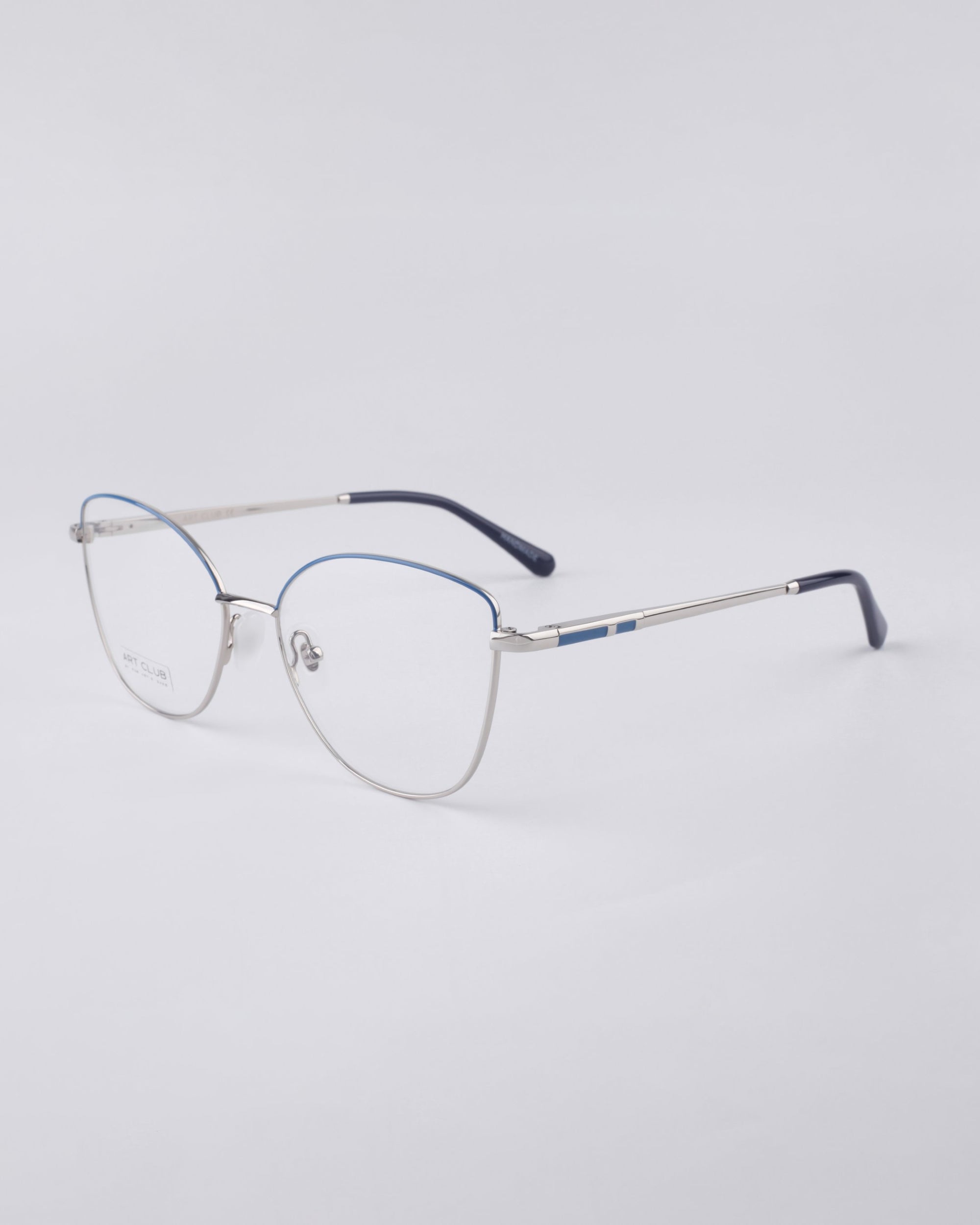 A pair of sleek, silver-framed For Art&#39;s Sake® eyeglasses with thin, metallic arms and blue-tipped earpieces rests on a plain white surface. These minimalist Greta frames feature clear lenses, crafted for sustainable luxury and modern aesthetics.