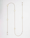 A long, delicate gold-plated necklace with a variety of small geometric charms including circles, squares, and a quatrefoil. This minimalistic piece also doubles as an elegant eyewear chain featuring pearl-like freshwater pearl embellishments. It is laid out against a plain white background. The Hampstead Glasses Chain by For Art's Sake® is the perfect blend of functionality and elegance.