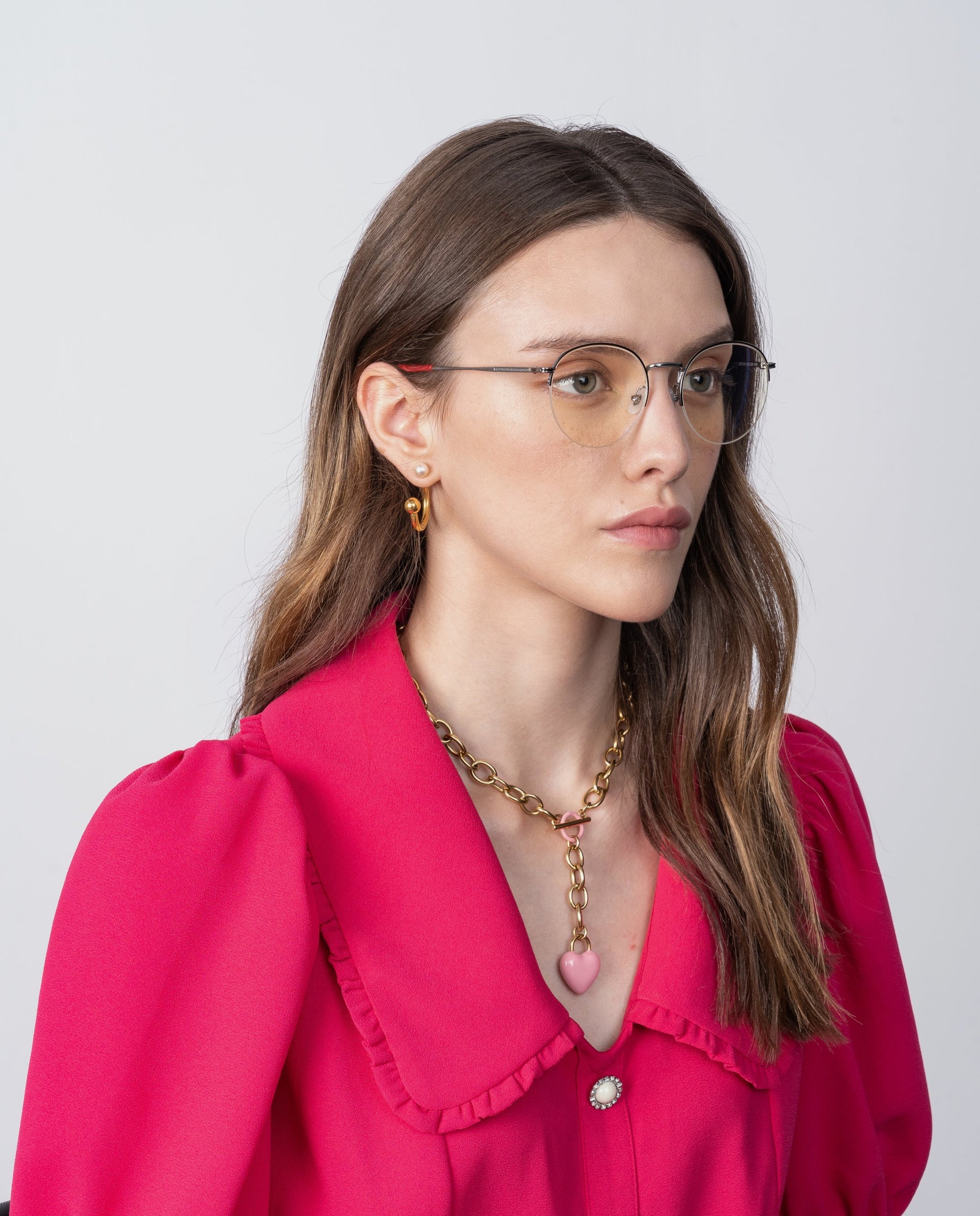 A woman with long brown hair, wearing Ivy glasses with blue light filter by For Art's Sake®, a bright pink blouse with puffed sleeves and a large collar, and a gold chain necklace with a heart-shaped pendant. She is looking slightly to the side against a plain background.