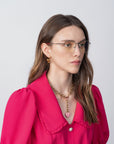 A woman with long brown hair, wearing Ivy glasses with blue light filter by For Art's Sake®, a bright pink blouse with puffed sleeves and a large collar, and a gold chain necklace with a heart-shaped pendant. She is looking slightly to the side against a plain background.