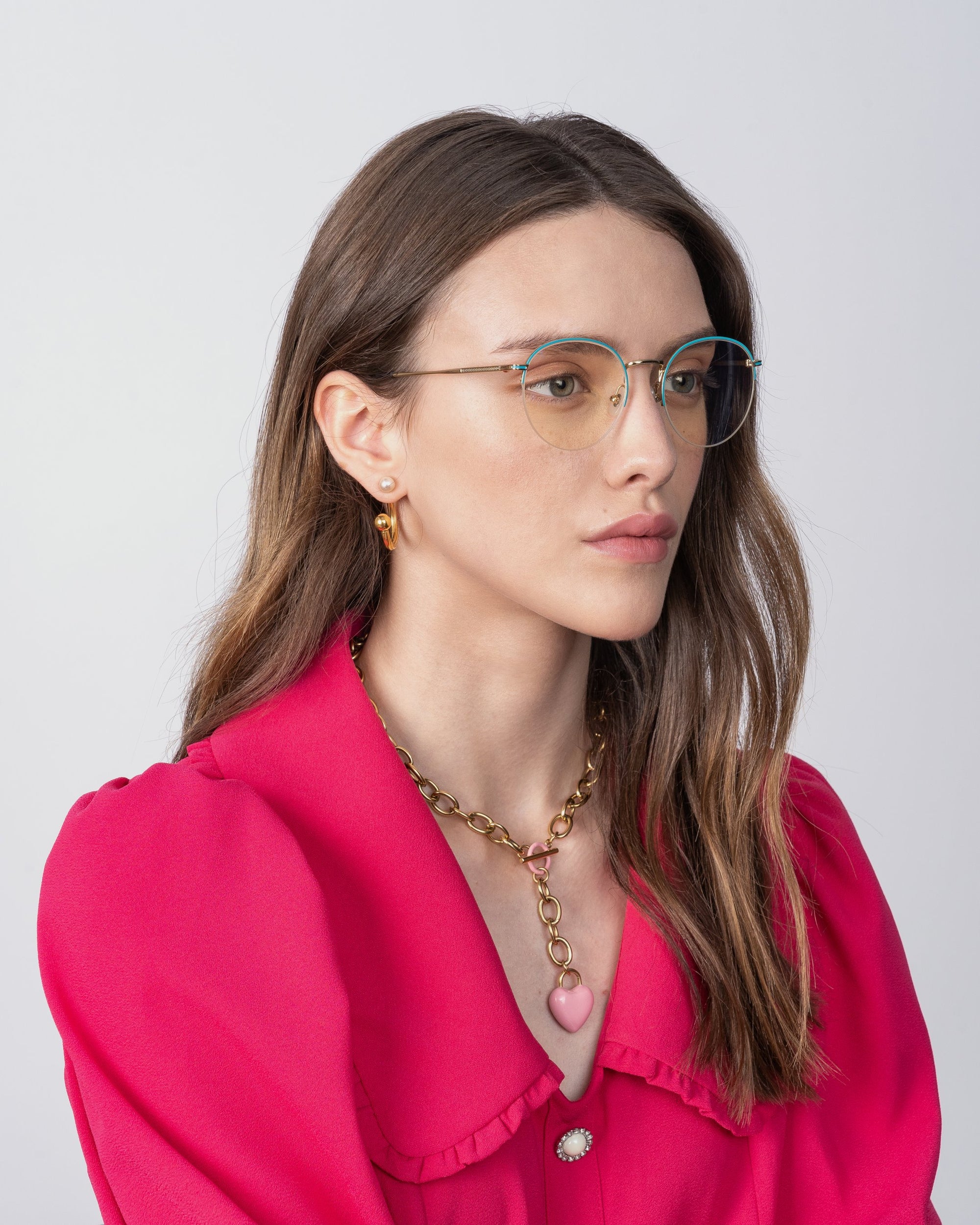 A woman with long, brown hair is wearing For Art's Sake® Ivy glasses with blue light filter lenses, a bright pink blouse with puff sleeves, and a gold necklace with a pink heart pendant. She is looking slightly to the side with a neutral expression against a plain white background.