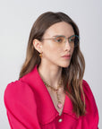 A woman with long, brown hair is wearing For Art's Sake® Ivy glasses with blue light filter lenses, a bright pink blouse with puff sleeves, and a gold necklace with a pink heart pendant. She is looking slightly to the side with a neutral expression against a plain white background.