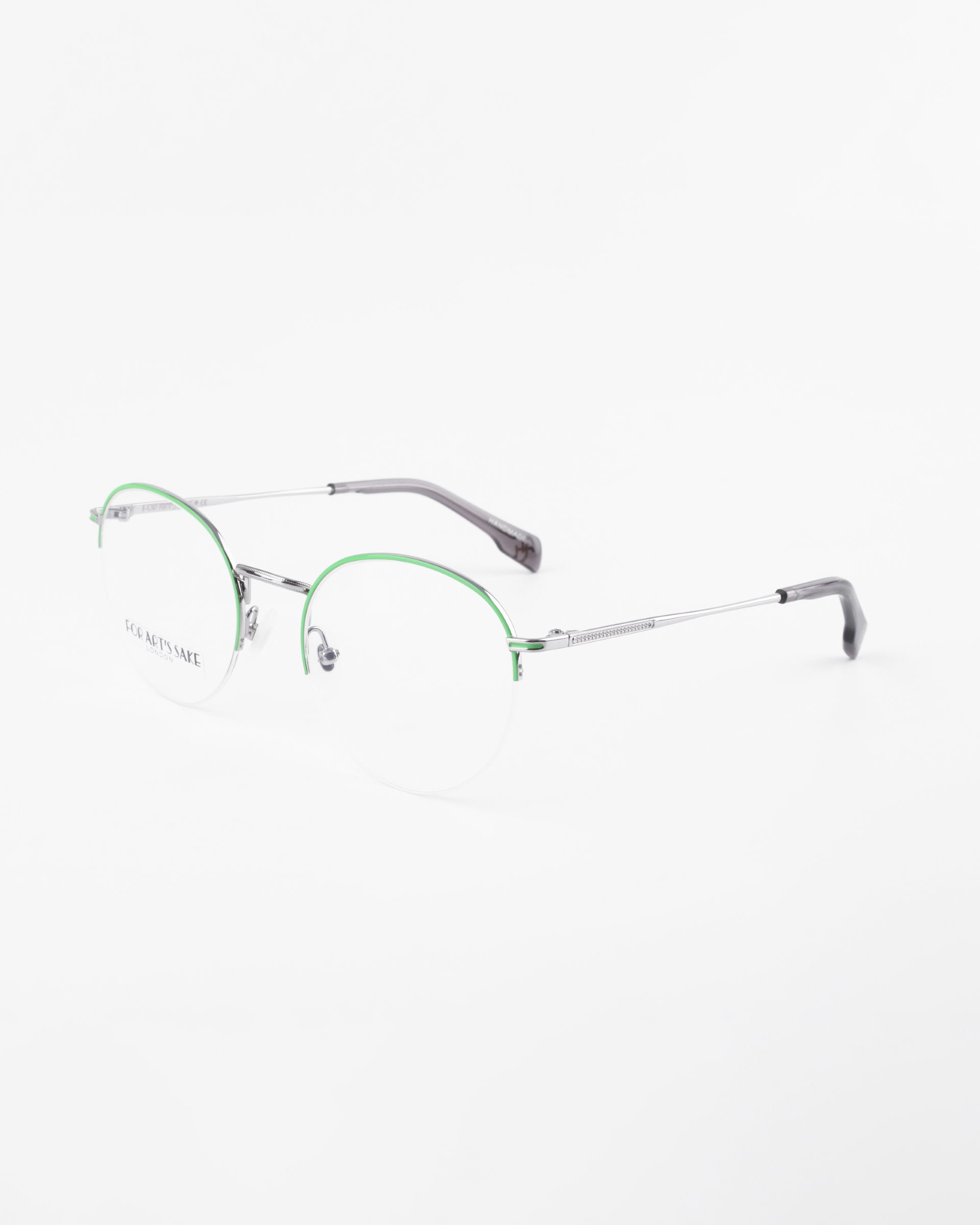A pair of For Art&#39;s Sake® Ivy eyeglasses with thin silver metal frames and green accents on the upper rims. The glasses, featuring clear round prescription lenses with a blue light filter, and dark-colored temple tips, are placed on a white background.