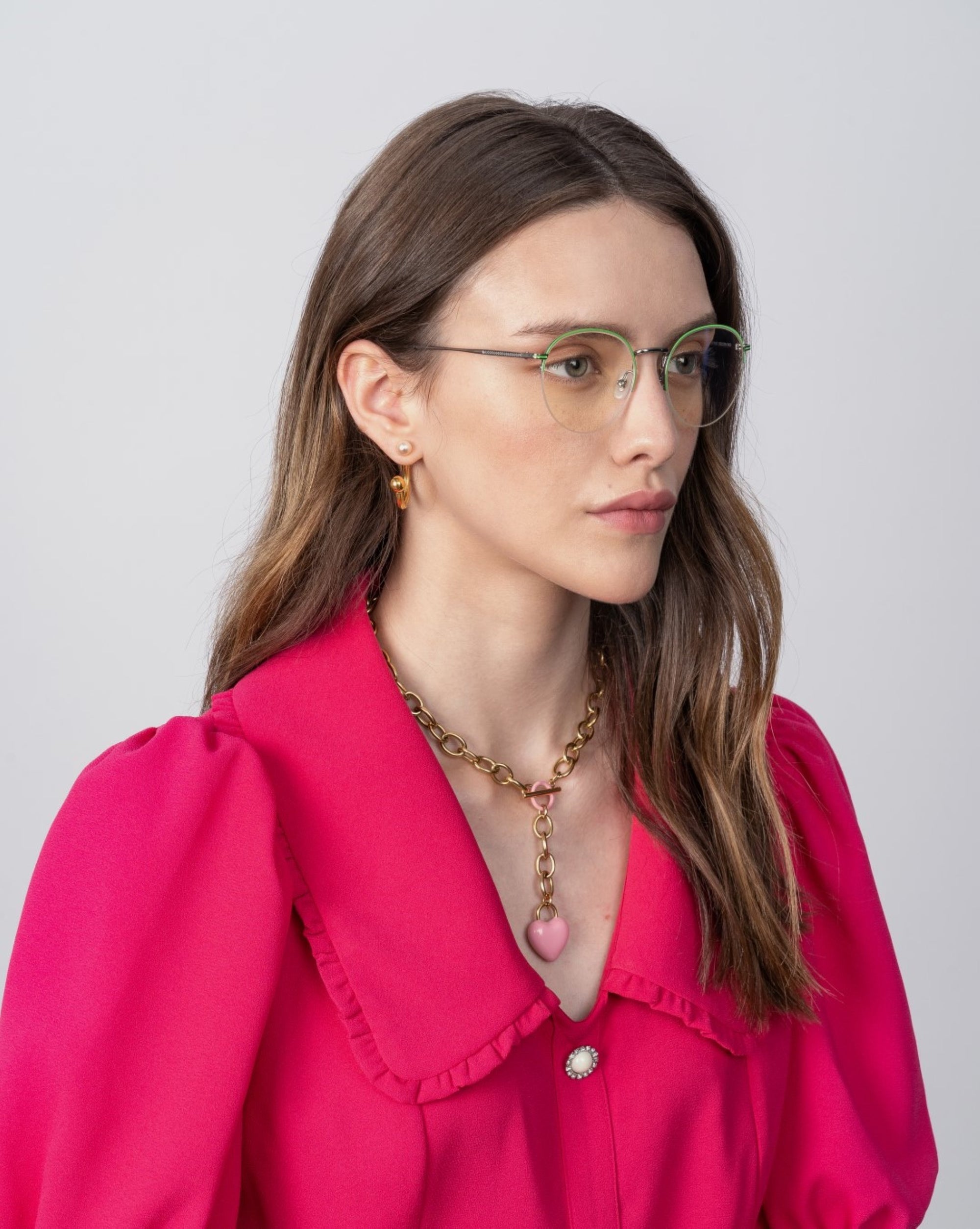 A person with long brown hair, wearing For Art&#39;s Sake® Ivy glasses with a blue light filter and a vibrant fuchsia blouse with a ruffled collar, looks to the left. They have a gold necklace with a heart pendant and small hoop earrings. The background is plain and light-colored.