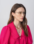 A person with long brown hair, wearing For Art's Sake® Ivy glasses with a blue light filter and a vibrant fuchsia blouse with a ruffled collar, looks to the left. They have a gold necklace with a heart pendant and small hoop earrings. The background is plain and light-colored.