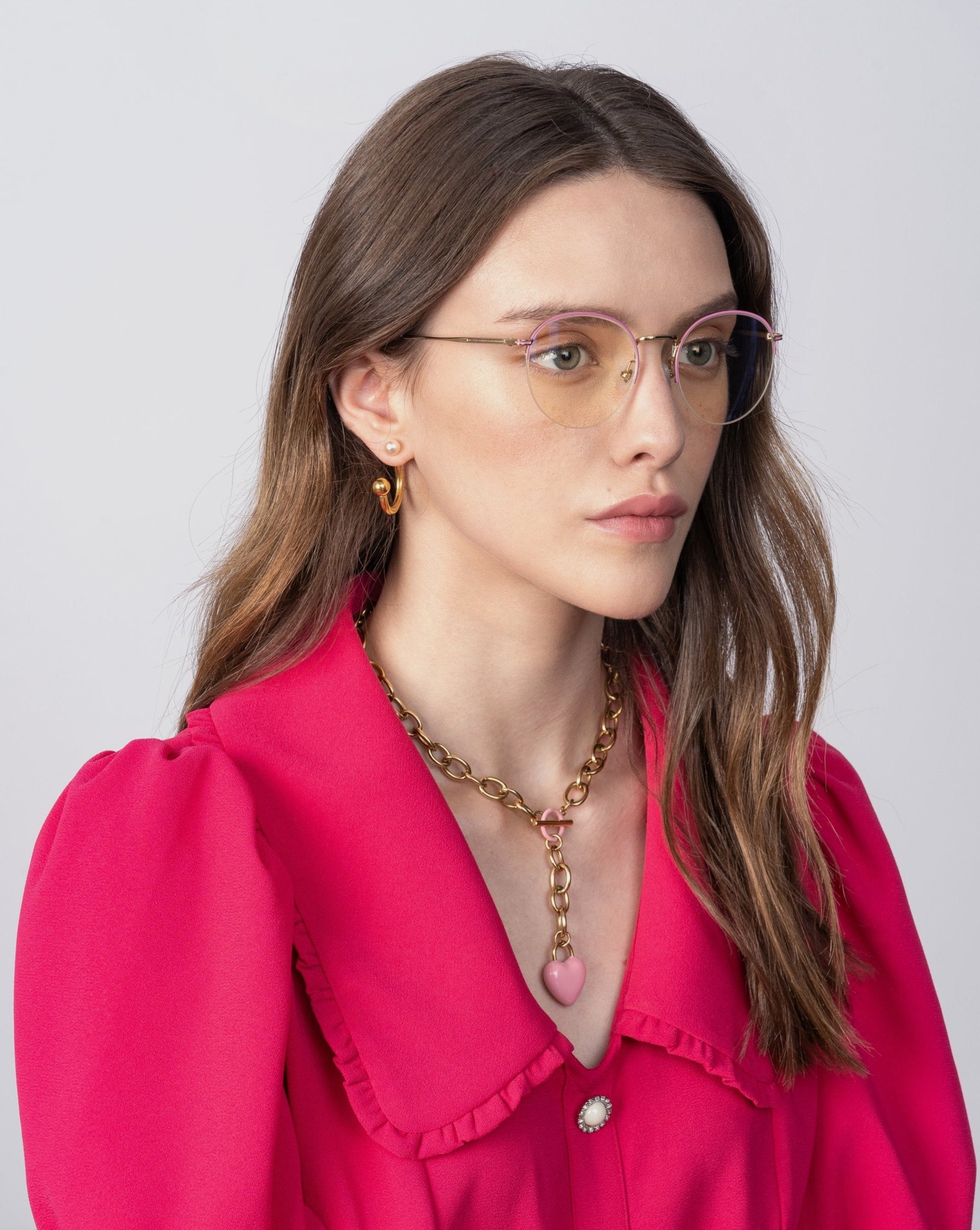 A woman with long brown hair wears a bright pink blouse with puffed sleeves and a red collar. She accessorizes with Ivy glasses by For Art&#39;s Sake® featuring prescription lenses, gold hoop earrings, and a chunky gold chain necklace with a pink heart pendant. The background is plain and light-colored.