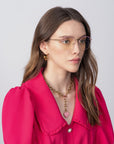 A woman with long brown hair wears a bright pink blouse with puffed sleeves and a red collar. She accessorizes with Ivy glasses by For Art's Sake® featuring prescription lenses, gold hoop earrings, and a chunky gold chain necklace with a pink heart pendant. The background is plain and light-colored.