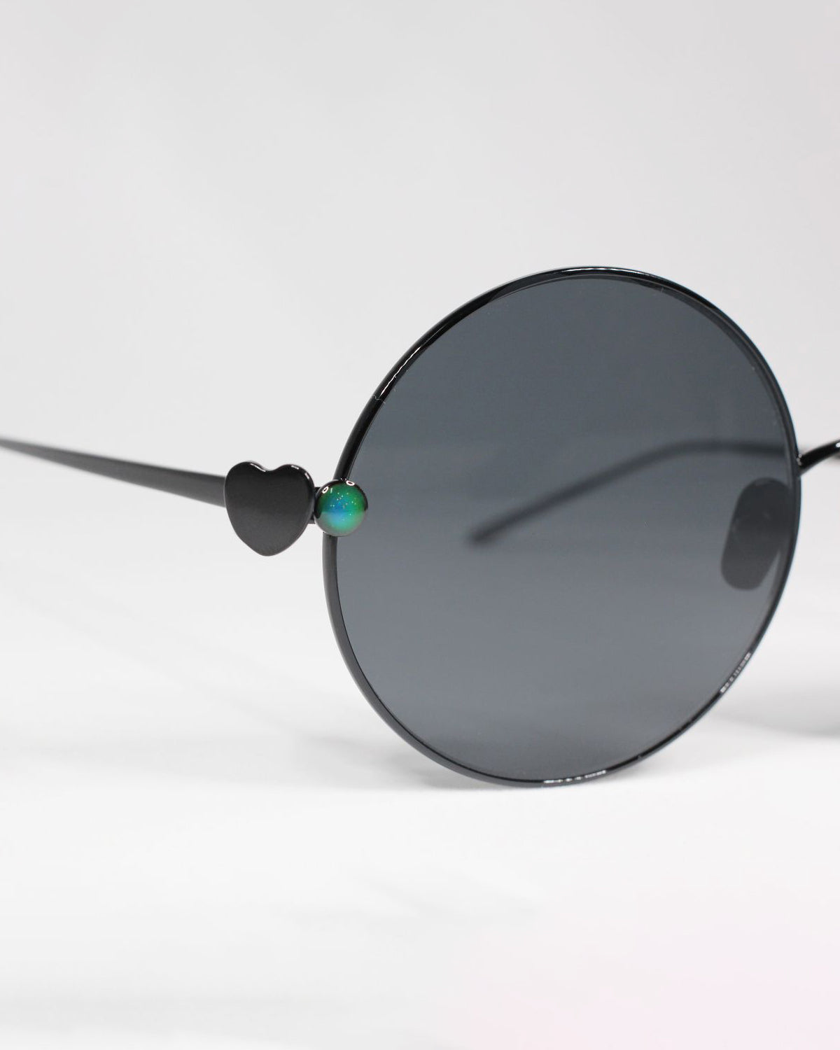 A pair of For Art&#39;s Sake® Love Story shades with round dark lenses and thin, stainless steel frames. The temples are adorned with a small heart detail and a single, multicolored gem on each side near the lenses. Offering UV protection, they come against a plain white background.