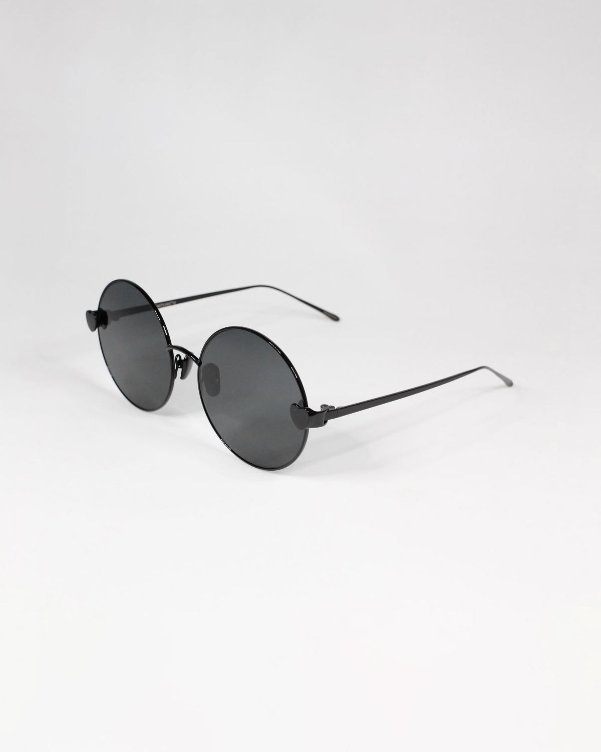 A pair of black round For Art&#39;s Sake® Love Story shades with thin stainless steel frames and dark lenses is placed on a plain white background. The design is minimalist and sleek, offering both style and UV protection.