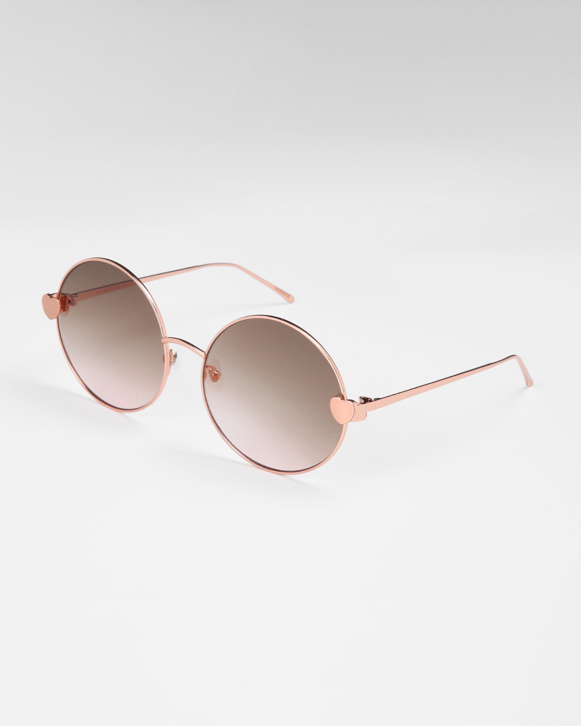 A pair of stylish For Art&#39;s Sake® Love Story shades with round sunglasses, featuring a rose gold frame and brown gradient lenses. The temples are sleek with thin arms, and there are heart-shaped accents where the lenses meet the frame. Equipped with jadestone nose pads for comfort and 100% UV protection. The background is plain white.