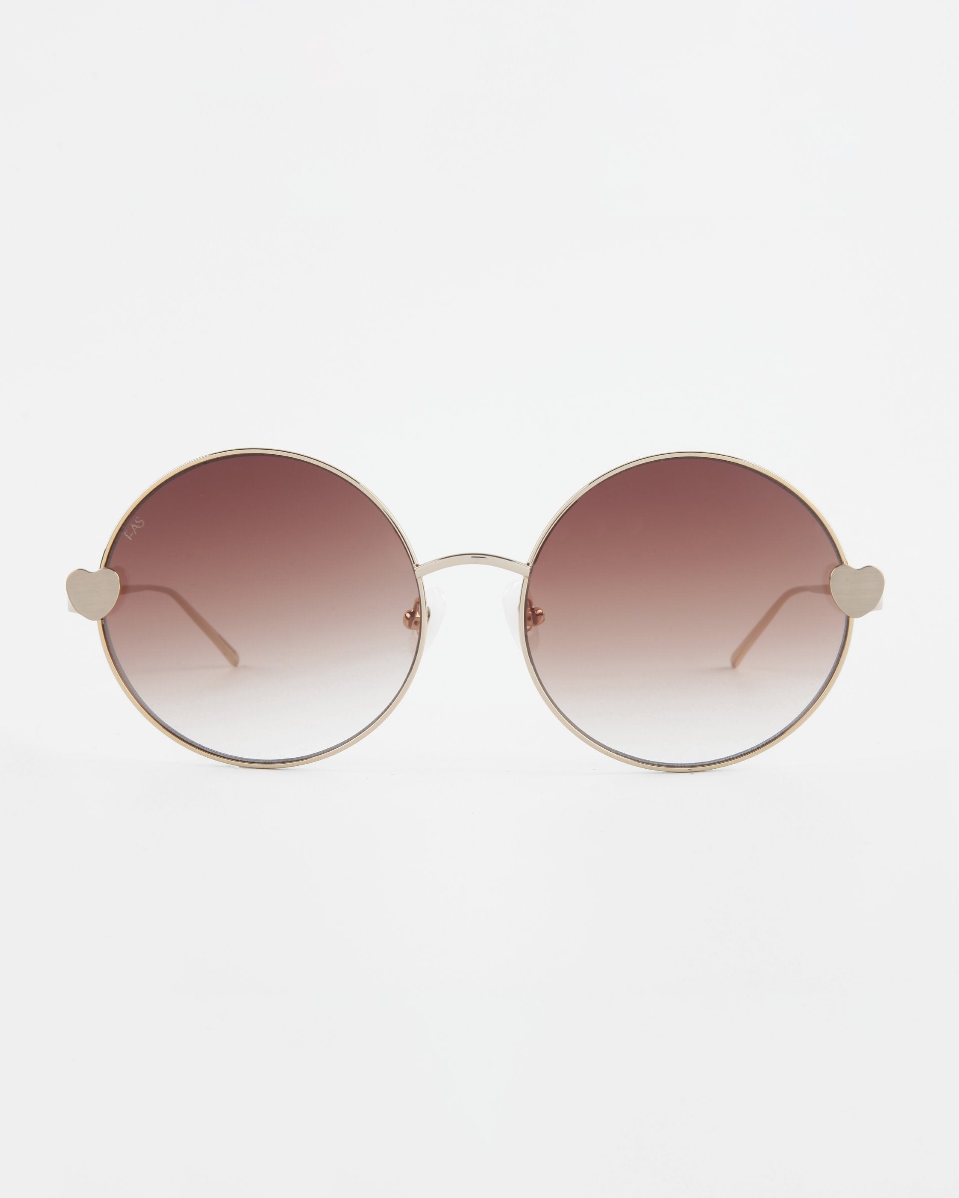 A pair of round For Art&#39;s Sake® Love Story shades with a thin gold frame and gradient brown lenses. These sunglasses feature small heart-shaped details at the hinges where the frames meet the lenses, jadestone nose pads for added comfort, and 100% UV protection. The background is plain white.