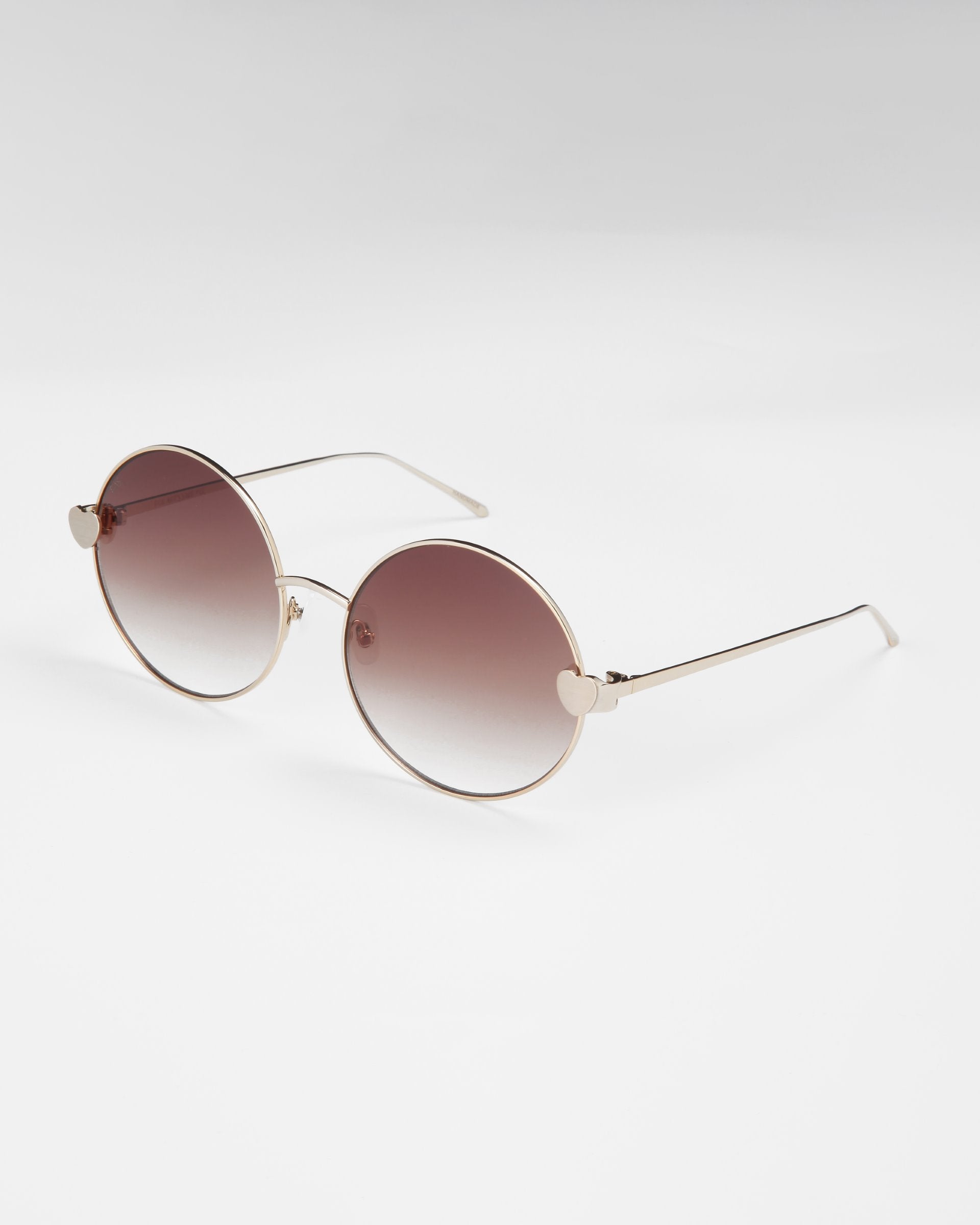 A pair of For Art&#39;s Sake® Love Story shades with round, gold-framed sunglasses featuring gradient brown lenses. The thin metal arms boast small heart-shaped accents near the hinges, and the jadestone nose pads ensure comfort. With 100% UV protection, these sunglasses rest elegantly on a light, neutral background.