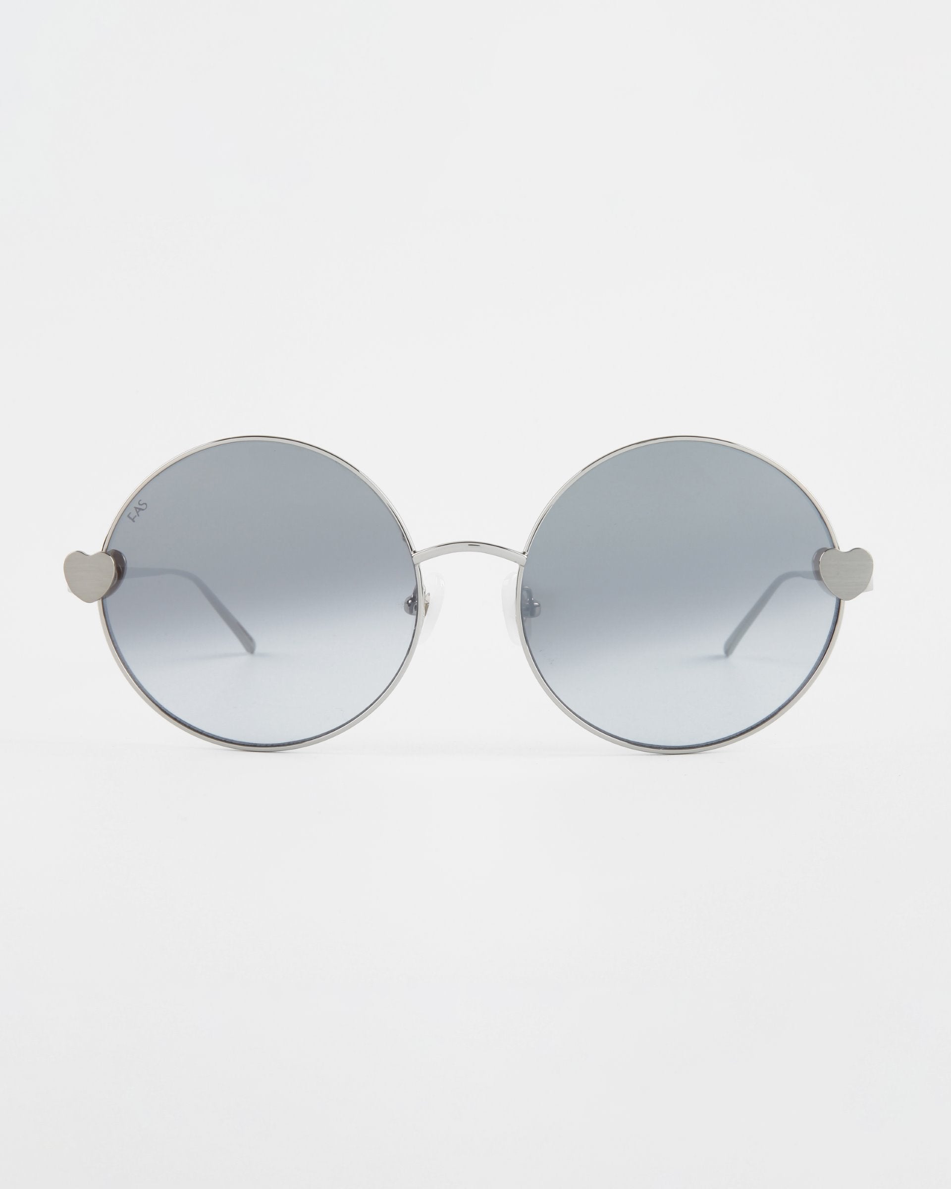 A pair of round For Art's Sake® Love Story shades with thin silver frames and grey-tinted lenses. The hinges on both sides are in the shape of small hearts, adding a unique and playful touch to the minimalist design. Equipped with jadestone nose pads, they also offer 100% UV protection against harmful rays. The background is plain white.