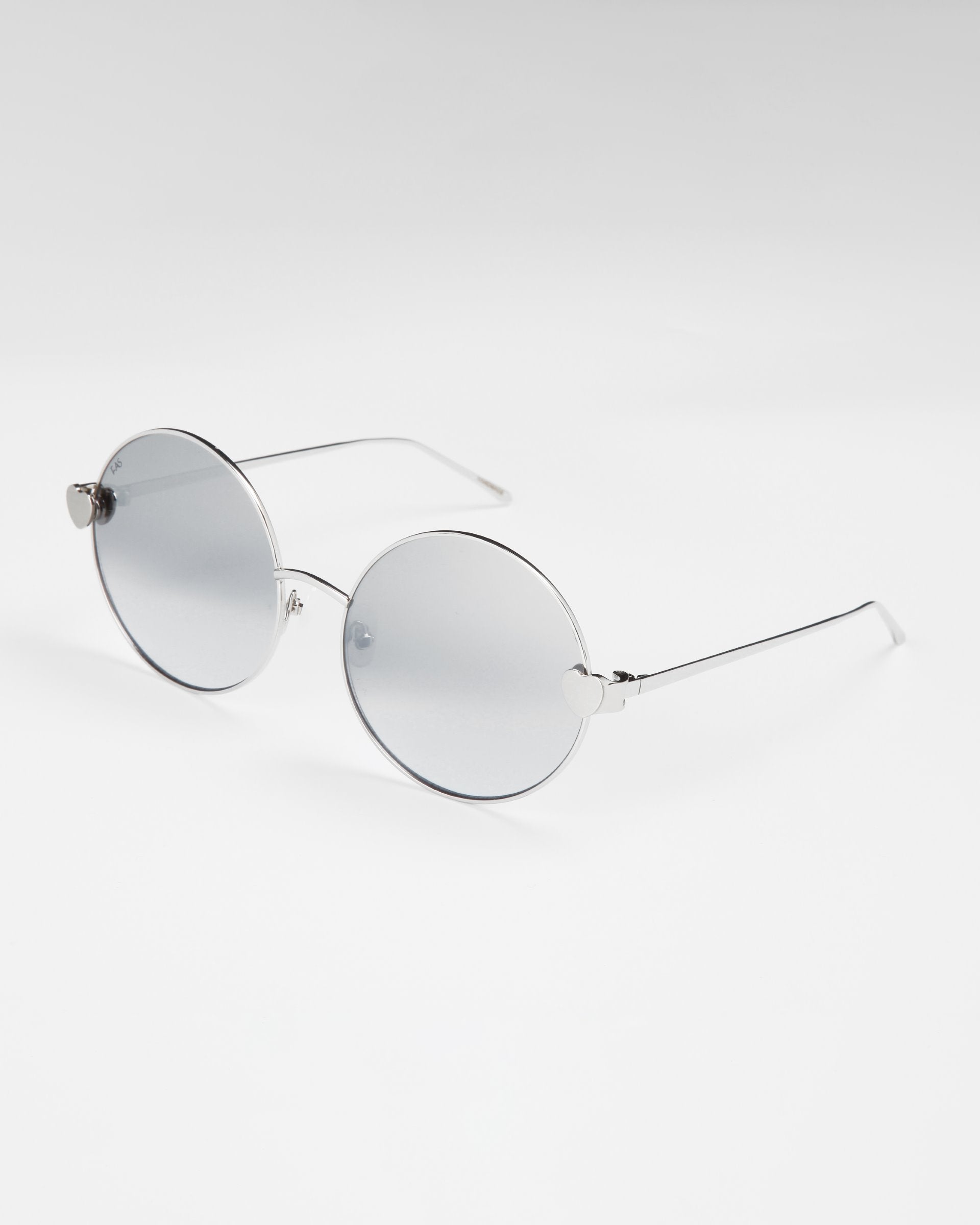A pair of round, silver-framed sunglasses with light gray lenses is set against a white background. The frames are thin and minimalist, with slightly curved temples and jadestone nose pads. These For Art&#39;s Sake® Love Story shades also offer 100% UV protection for both style and safety.