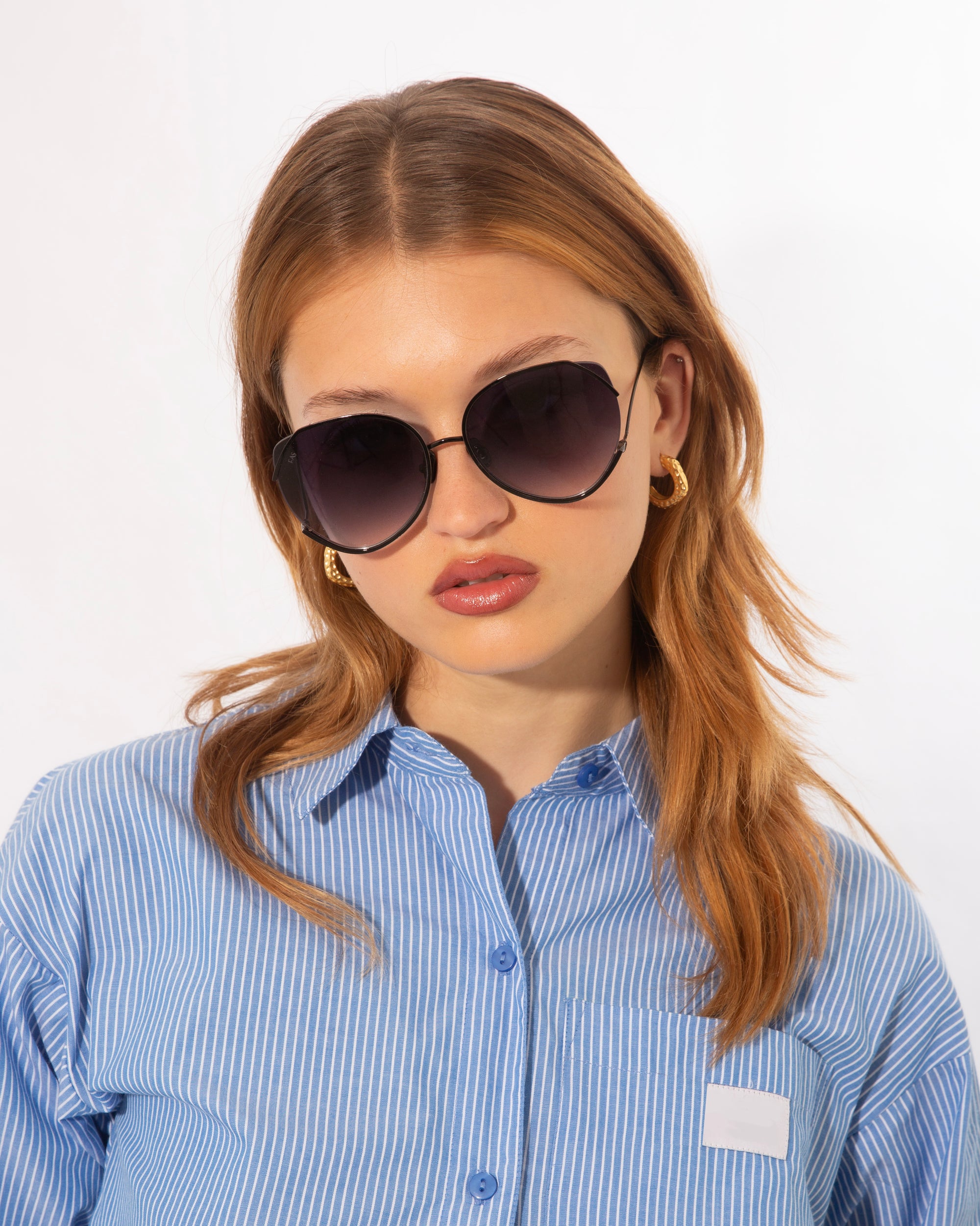A woman wearing large, round, dark sunglasses with ultralightweight lenses and jadestone nose pads gazes into the camera. She has long, light brown hair and is dressed in a blue and white striped shirt. The background is plain white, highlighting her stylish For Art's Sake® Wonderland accessories offering 100% UV protection.